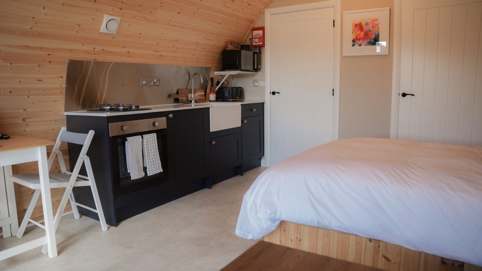 The cozy wooden cabin interior at Partridge Retreat features a small kitchen area with a stove, oven, and sink. To the right is a bed with white bedding, while to the left sits a small dining table and chair. Decorative art adorns the walls, enhancing this peaceful hideaway's charm.