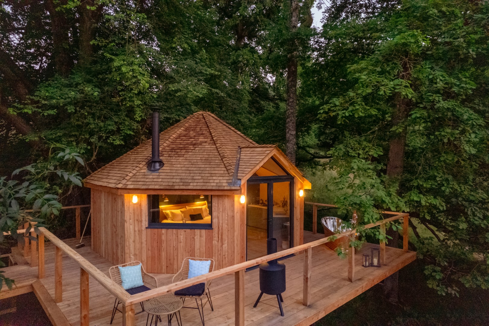 ​​​​Cosy wooden cabin with a deck nestled among trees, offering a secluded getaway location with outdoor seating and warm interior lighting.