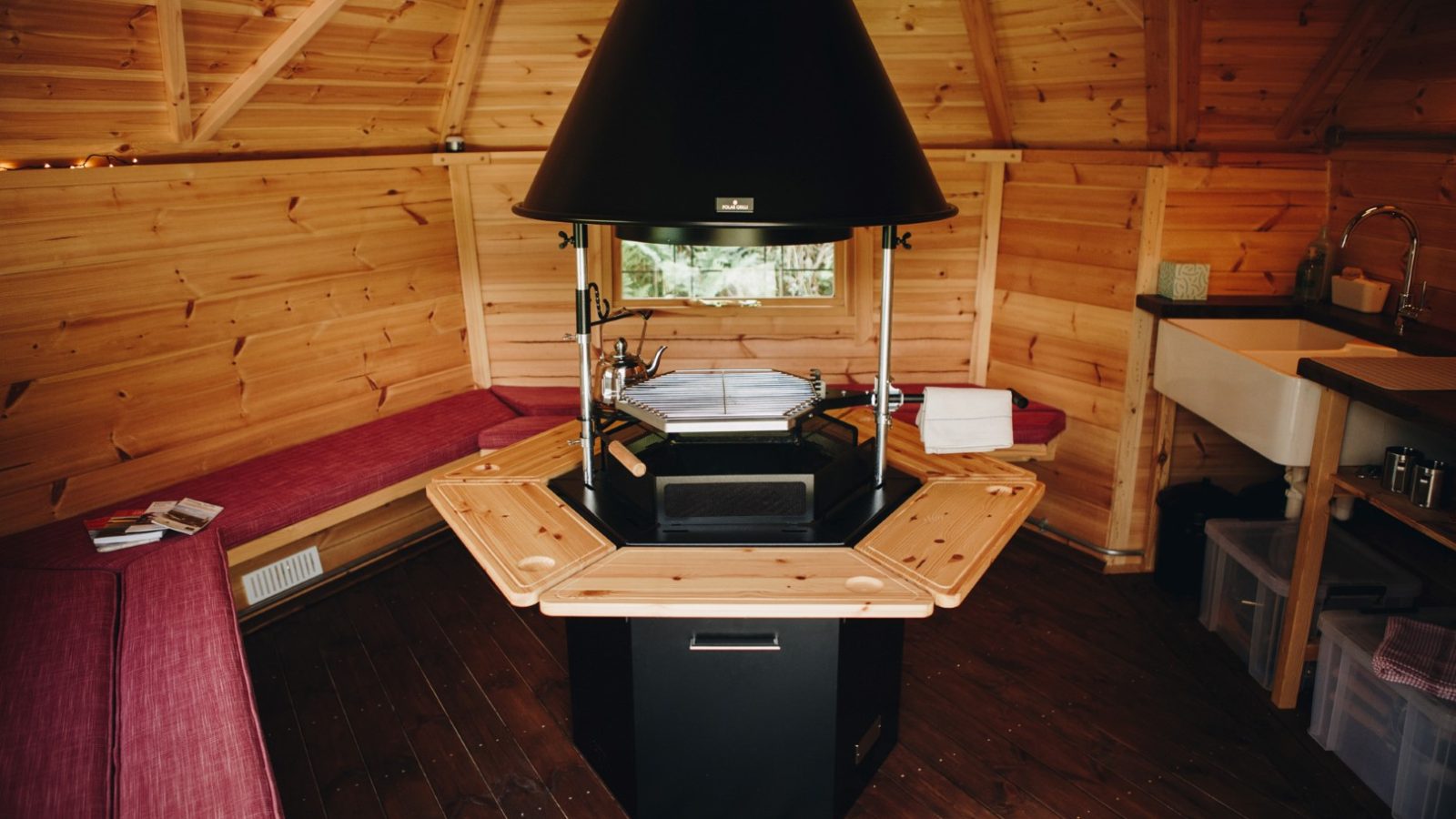 The cozy wooden cabin interior at Little Menherion features a central hexagonal grill surrounded by benches. Walls and ceiling boast light wood, while a small sink sits in the corner. Natural light pours through the window, crafting a warm and inviting atmosphere.