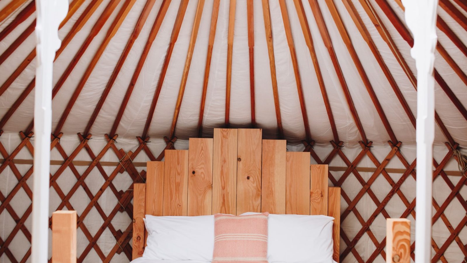 The cozy interior of Little Menherion's yurt features wooden lattice walls and radial ceiling beams. A bed with white sheets and a single pink pillow is centered, framed by two wooden posts. The atmosphere is warm and inviting.