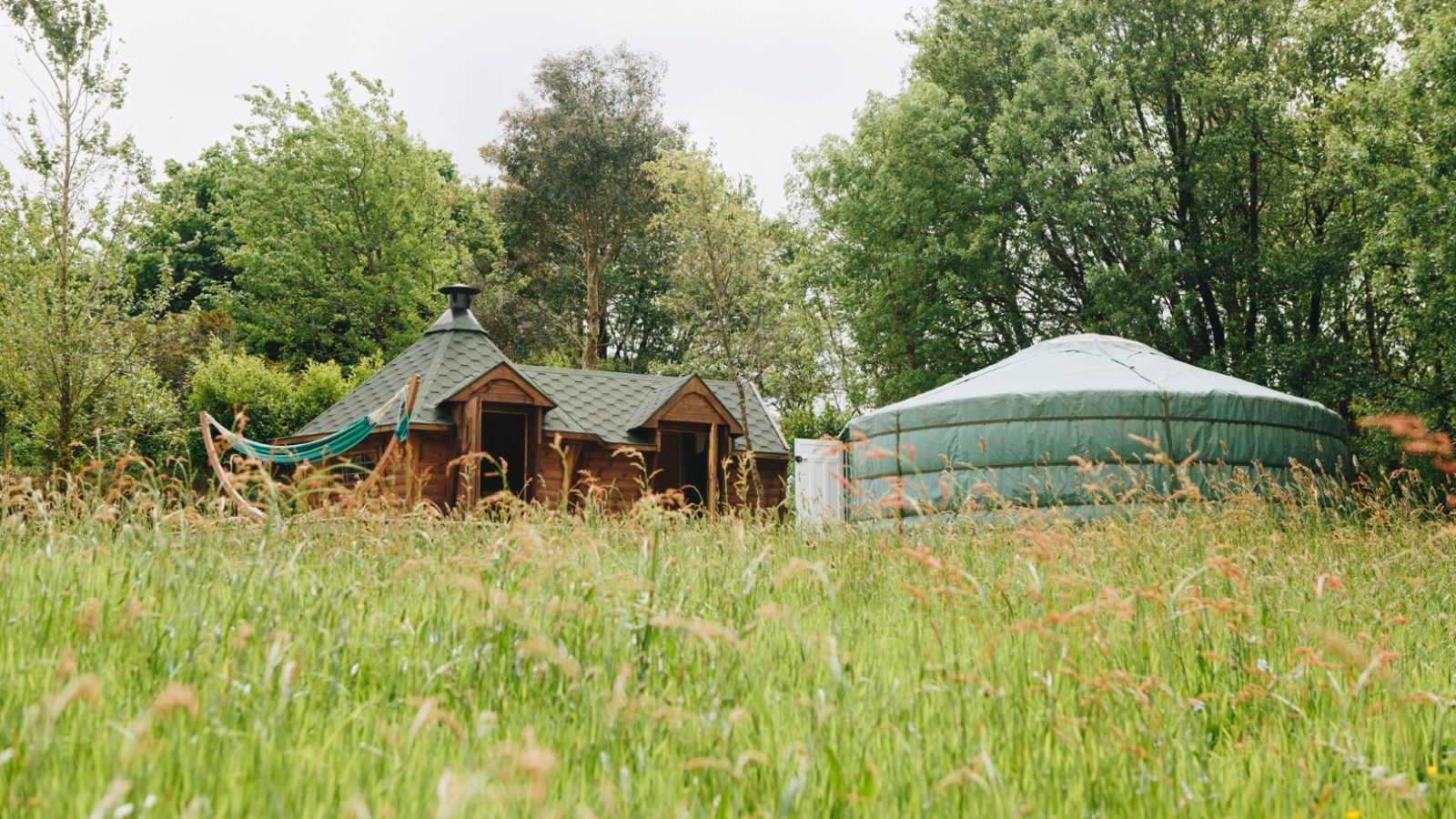 A lush green field with a wooden cabin and a circular tent graces Little Menherion, surrounded by tall grass and trees. The overcast sky adds to the serene, rustic atmosphere of this enchanting locale.