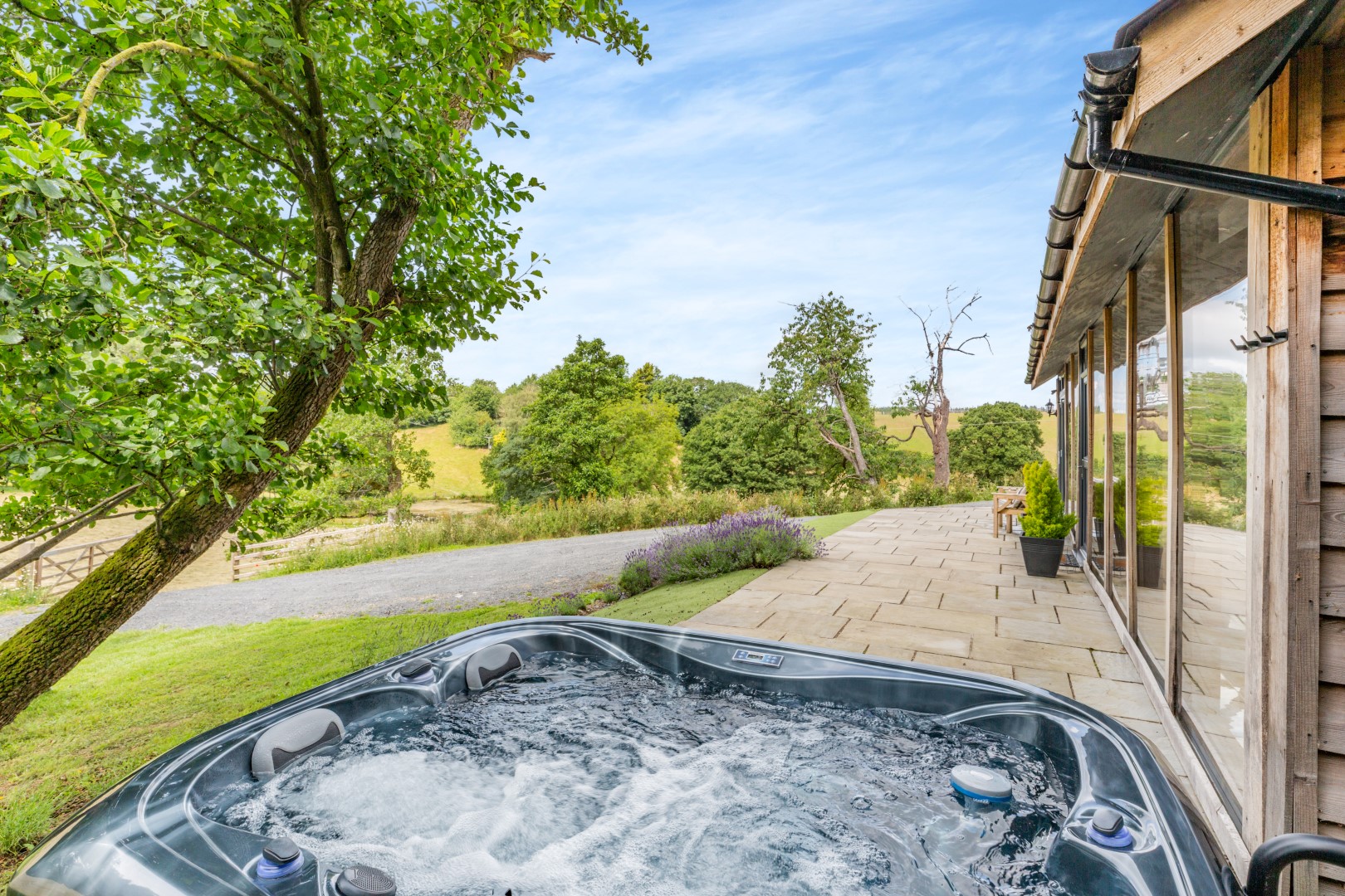 Experience a luxury wellness retreat with an outdoor hot tub set on a stone patio, offering a scenic view of the lush green landscape and a majestic tree under the clear blue sky.