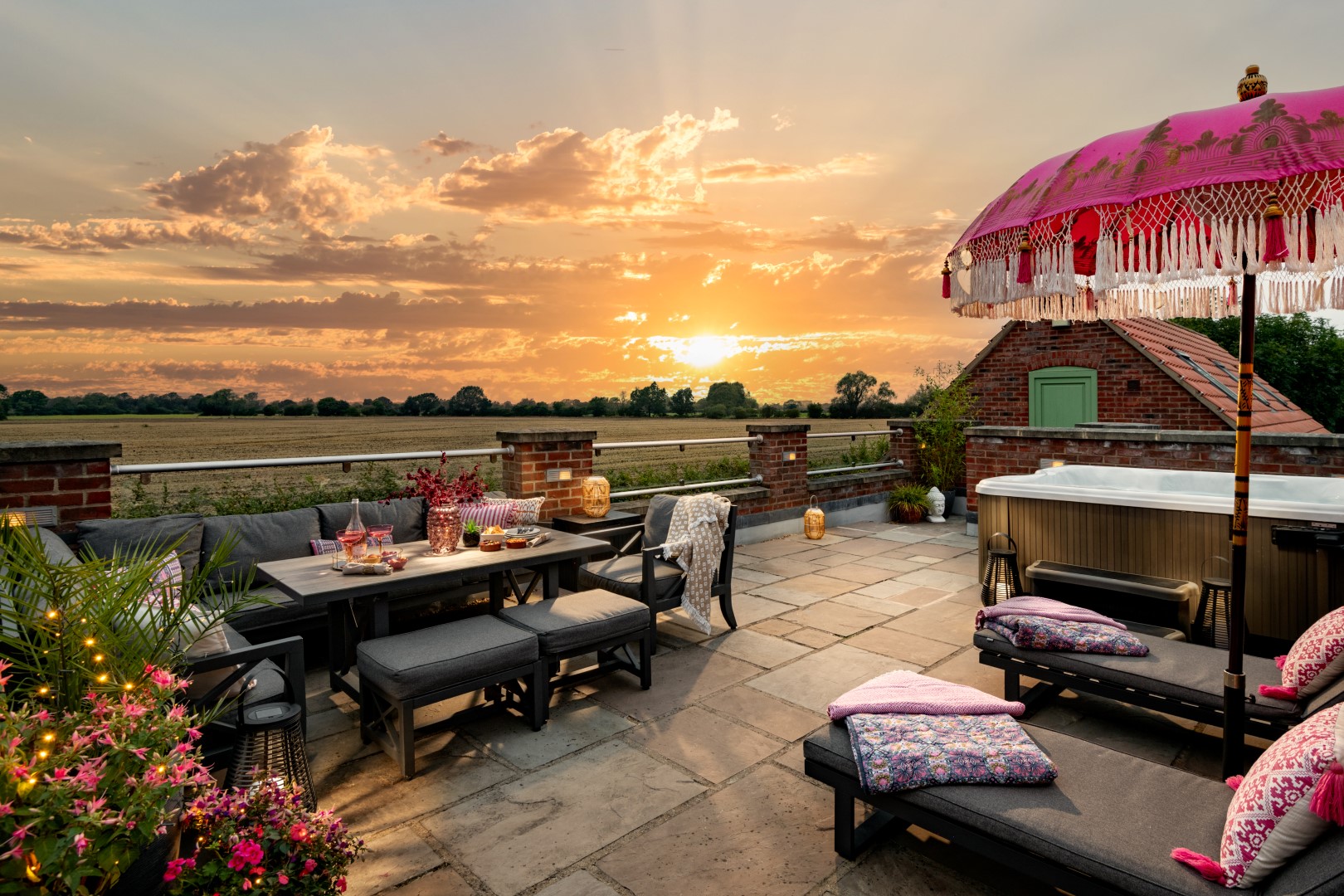 Rooftop patio with seating, a colourful parasol, and a hot tub welcoming sunset views. Fields and a house are visible in the background, offering the perfect setting for a luxury wellness retreat with family and friends in the UK.