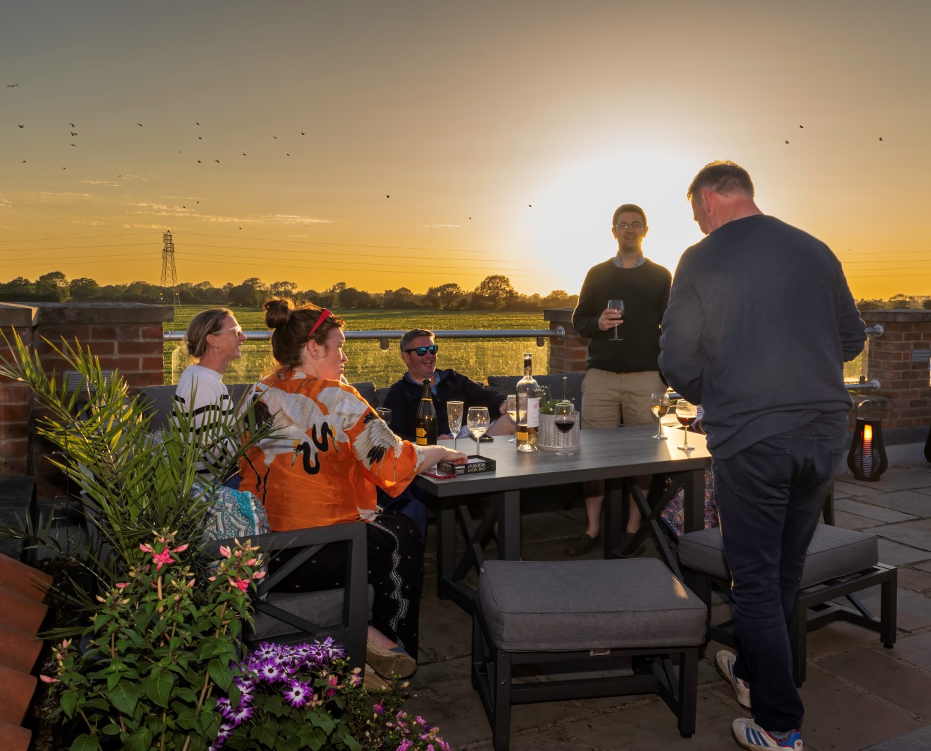 Five people gathered around an outdoor table at sunset, enjoying drinks and conversation during a summer holiday, with a scenic view in the background.