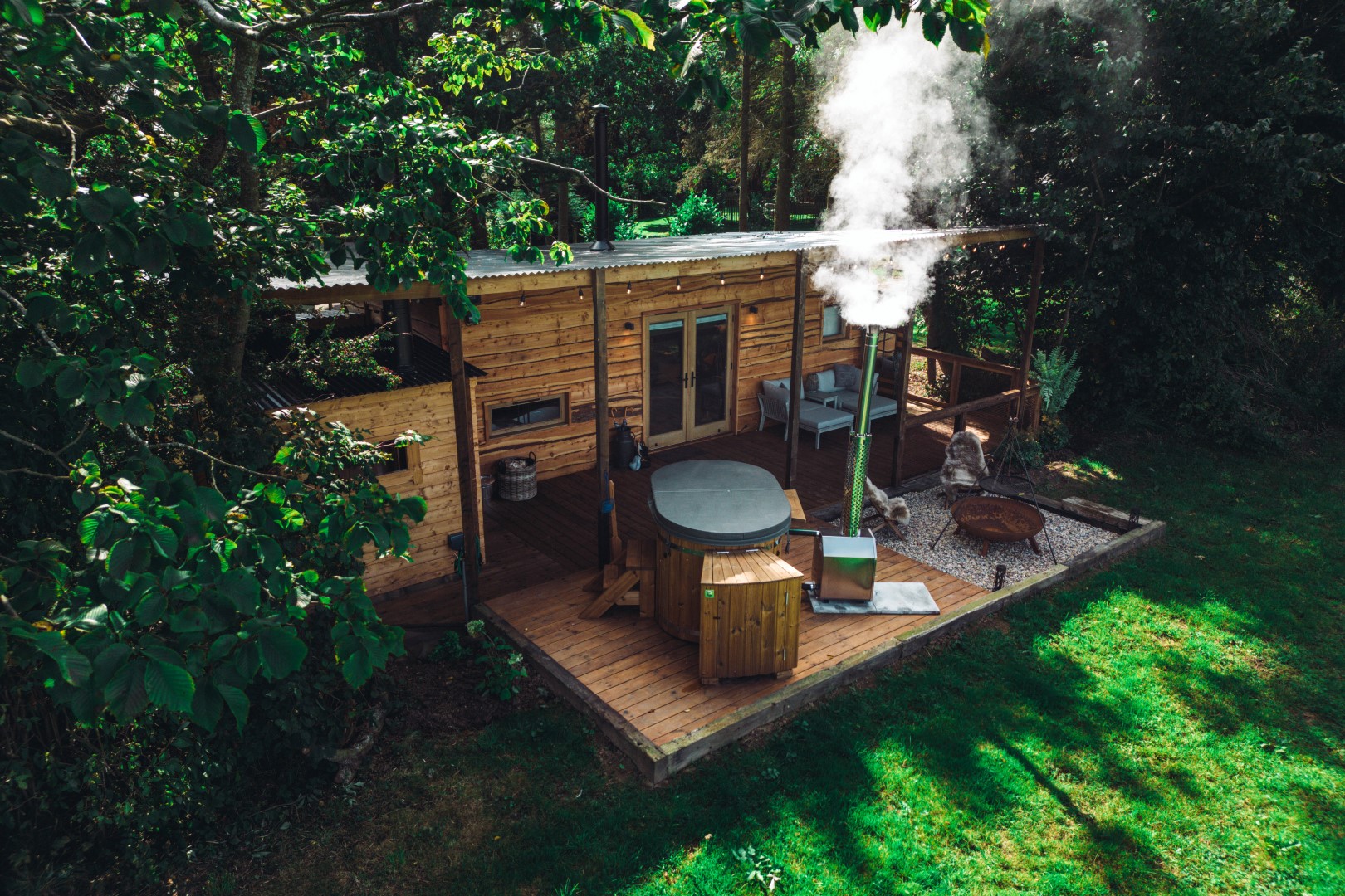 A luxury wooden cabin with a deck, hot tub, and quaint chimney emitting smoke, perfectly nestled amidst lush greenery for a serene wellness retreat in the UK.