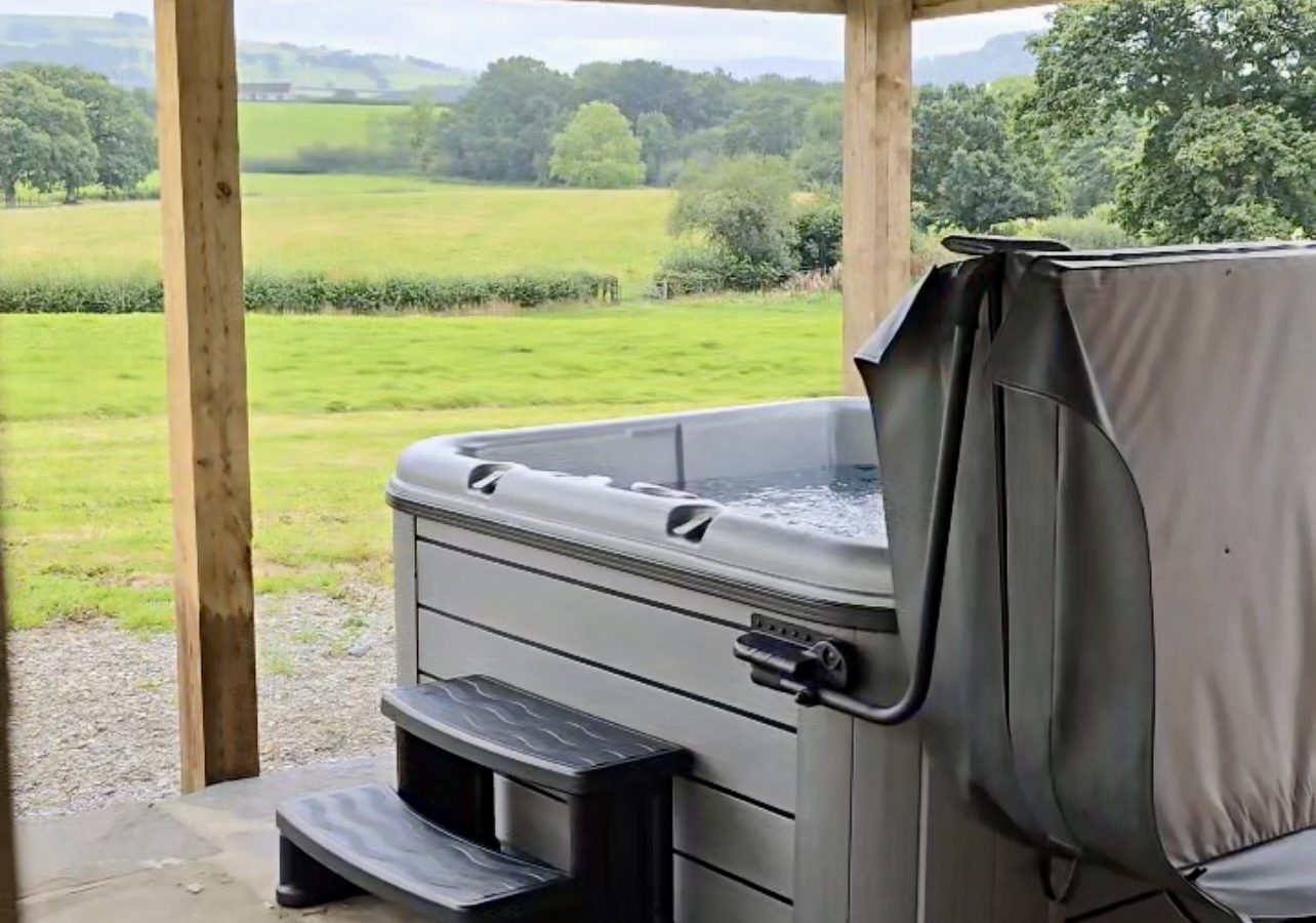 A covered outdoor hot tub is set on a stone patio with a view of lush green fields and the distant Black Mountain. The wooden roof structure adorned with string lights, and two steps lead up to the tub. The scene at Black Mountain Escapes is tranquil and inviting.