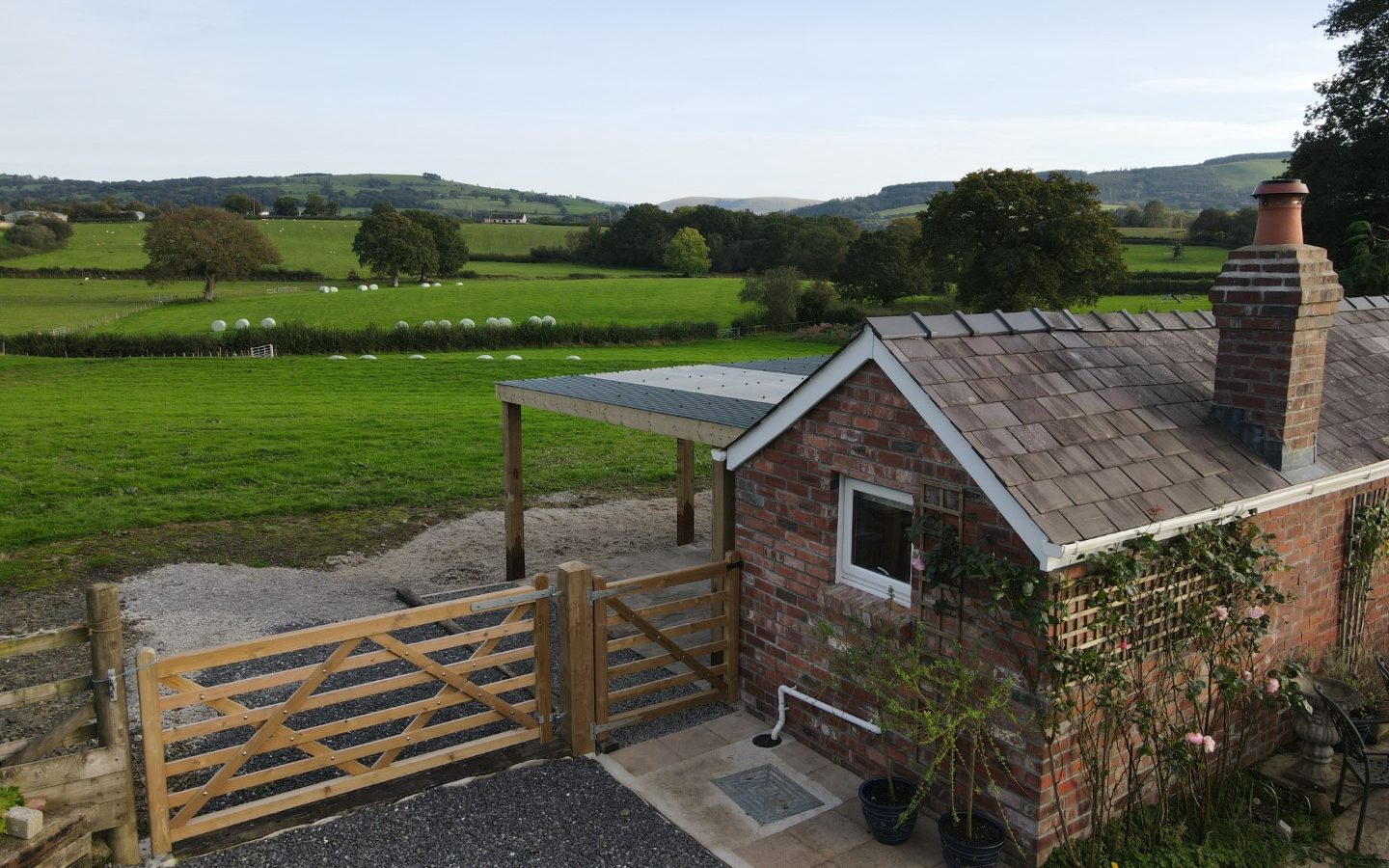 A small brick cottage with a tiled roof, part of Golchdy at Black Mountain Escapes, sits beside a wooden gate in a rural landscape. The scene is framed by green fields, trees, and rolling hills under a clear blue sky. A charming pergola is attached to the cottage.