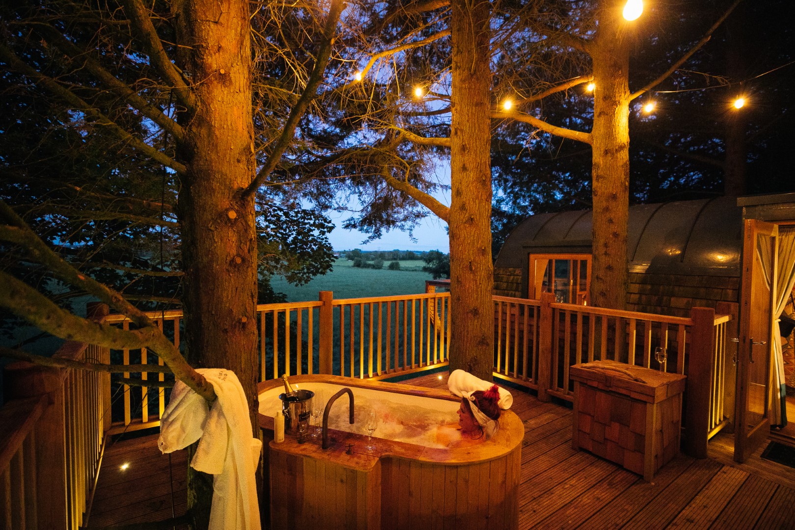 Experience a touch of luxury at this wellness retreat in the UK, featuring an outdoor wooden bathtub on a deck surrounded by trees. String lights twinkle overhead, with a serene field adding to the tranquil ambiance.