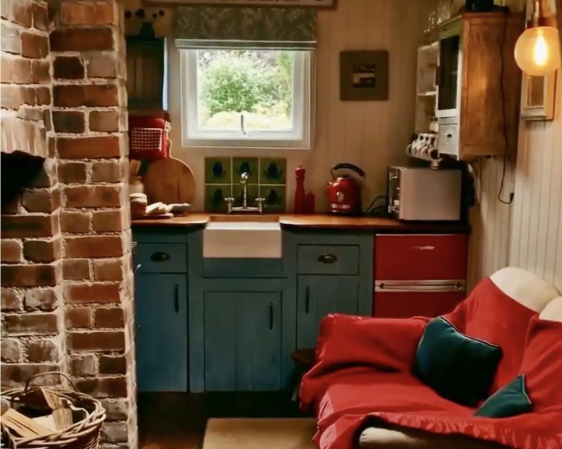 Cozy rustic kitchen by Y Golchdy with a small window above a wooden countertop, surrounded by brick and wood paneling. A red fridge and microwave rest on cabinets, while a beige loveseat with red blankets and pillows offers comfort in the foreground—perfect for Black Mountain Escapes.