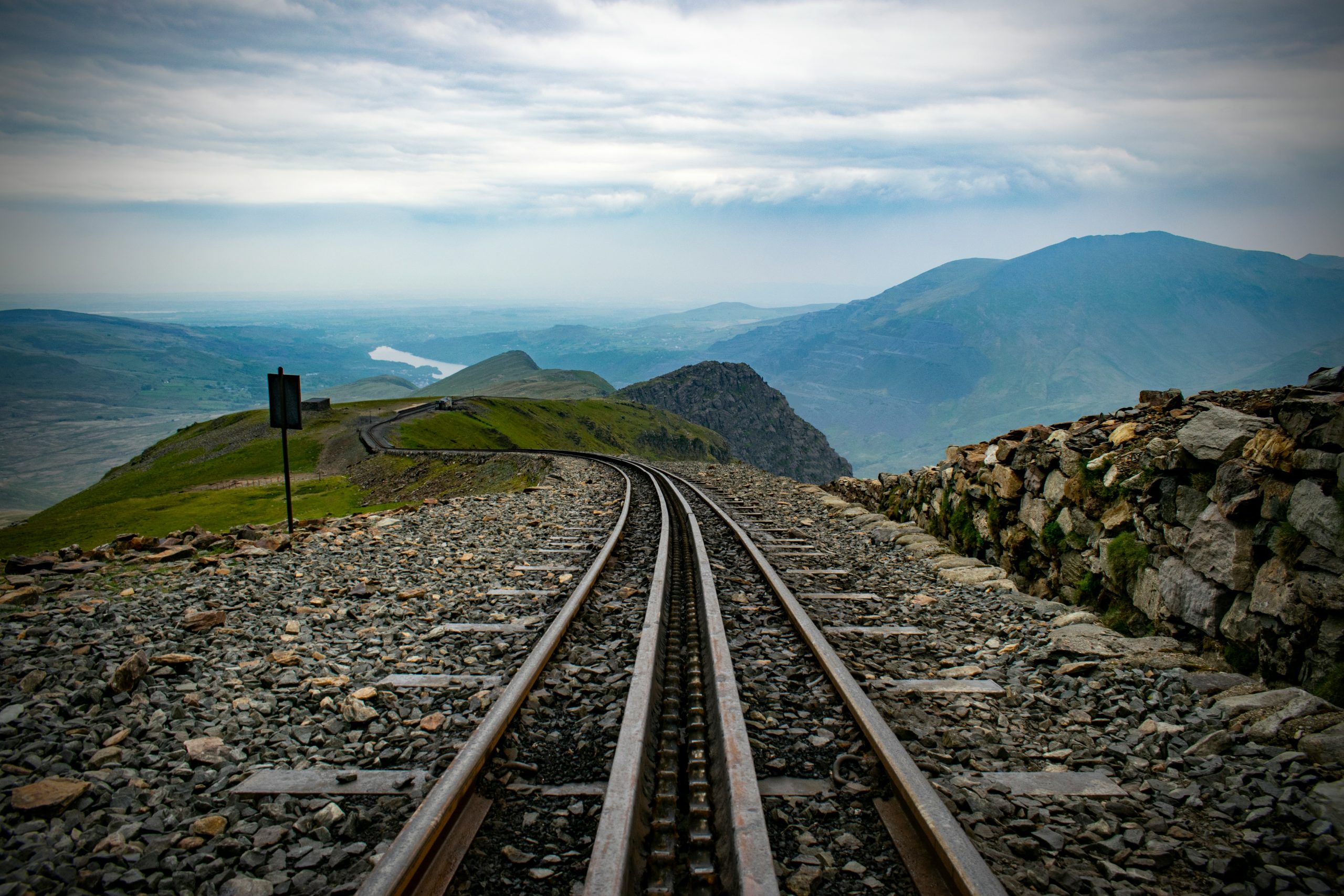 Railroad tracks lead through a mountainous landscape in North Wales under a cloudy sky, with stone walls lining the path, making it one of the enchanting travel destinations for tourism.