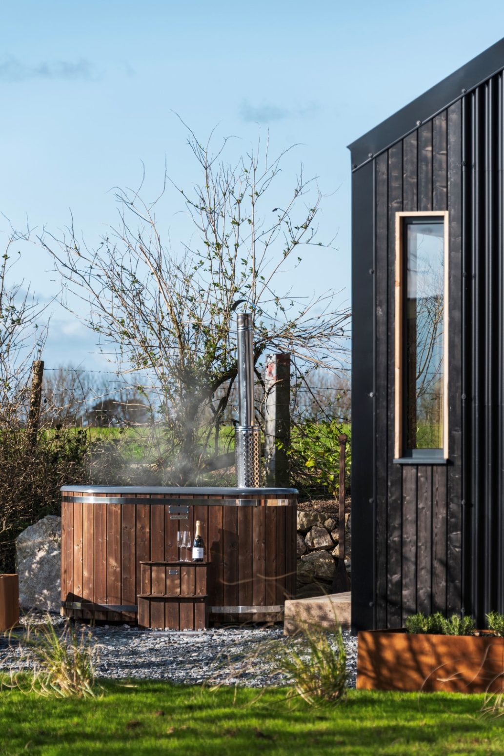 A wooden hot tub with steam gently rising sits beside a sleek, modern black building, surrounded by lush trees and grass. Perfect for showcasing tranquility and elegance, this scene is ideal for a home page that embodies relaxed luxury.