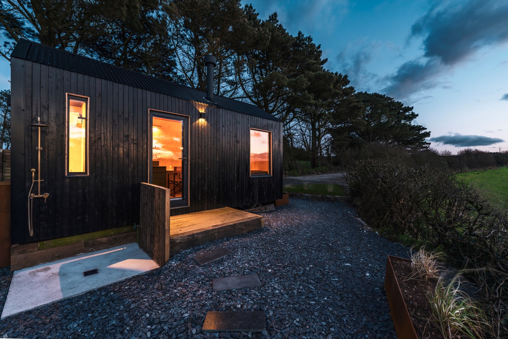 A small black wooden cabin with a lit interior stands on a gravel path, surrounded by trees and grass at dusk, offering the perfect escape for those seeking tranquil Coastal Cornwall breaks in the UK.