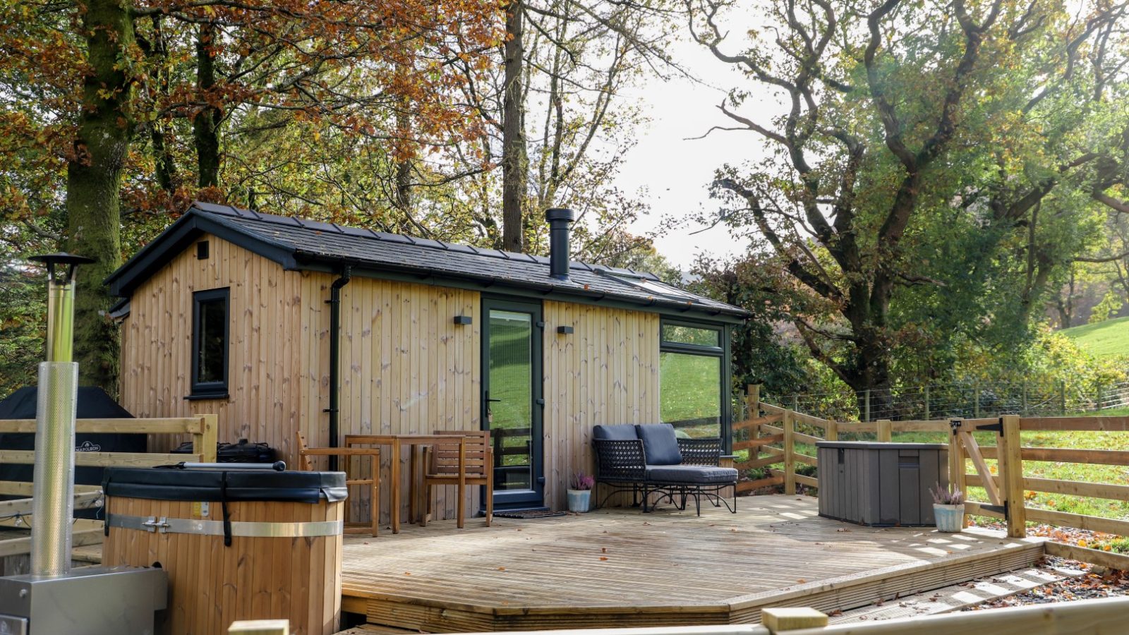 Wooden cabin with large windows and a deck featuring outdoor seating and a hot tub, surrounded by trees and a fenced yard—the perfect retreat for your Pennant holidays.