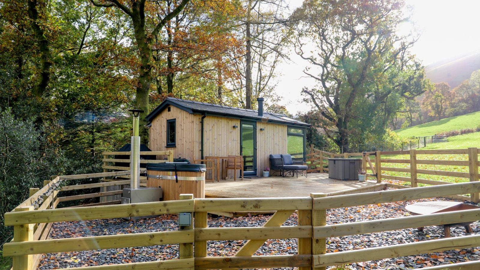 Experience the charm of a small wooden cabin at Gwyliau Pennant Holidays. Nestled amidst trees and a lush grassy landscape, this serene getaway features a fenced patio with outdoor seating and a hot tub, perfect for a relaxing holiday escape.