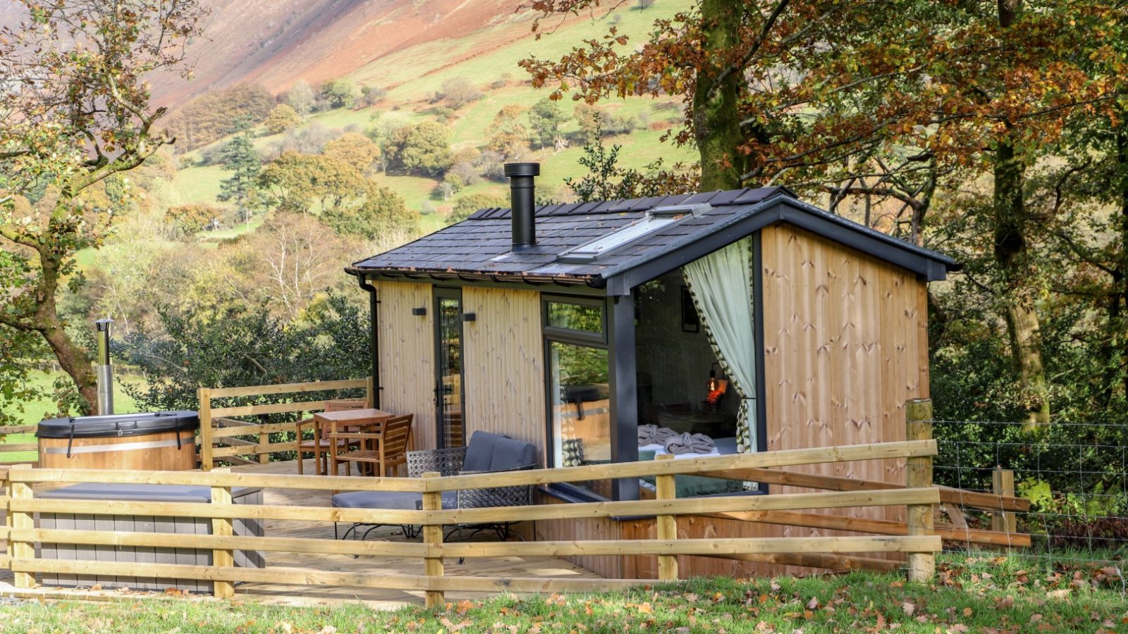 Escape to a small wooden cabin by Gwyliau Pennant Holidays, featuring a cozy deck with outdoor seating. Nestled among nearby trees, it offers serene views of the rural landscape and rolling hills.