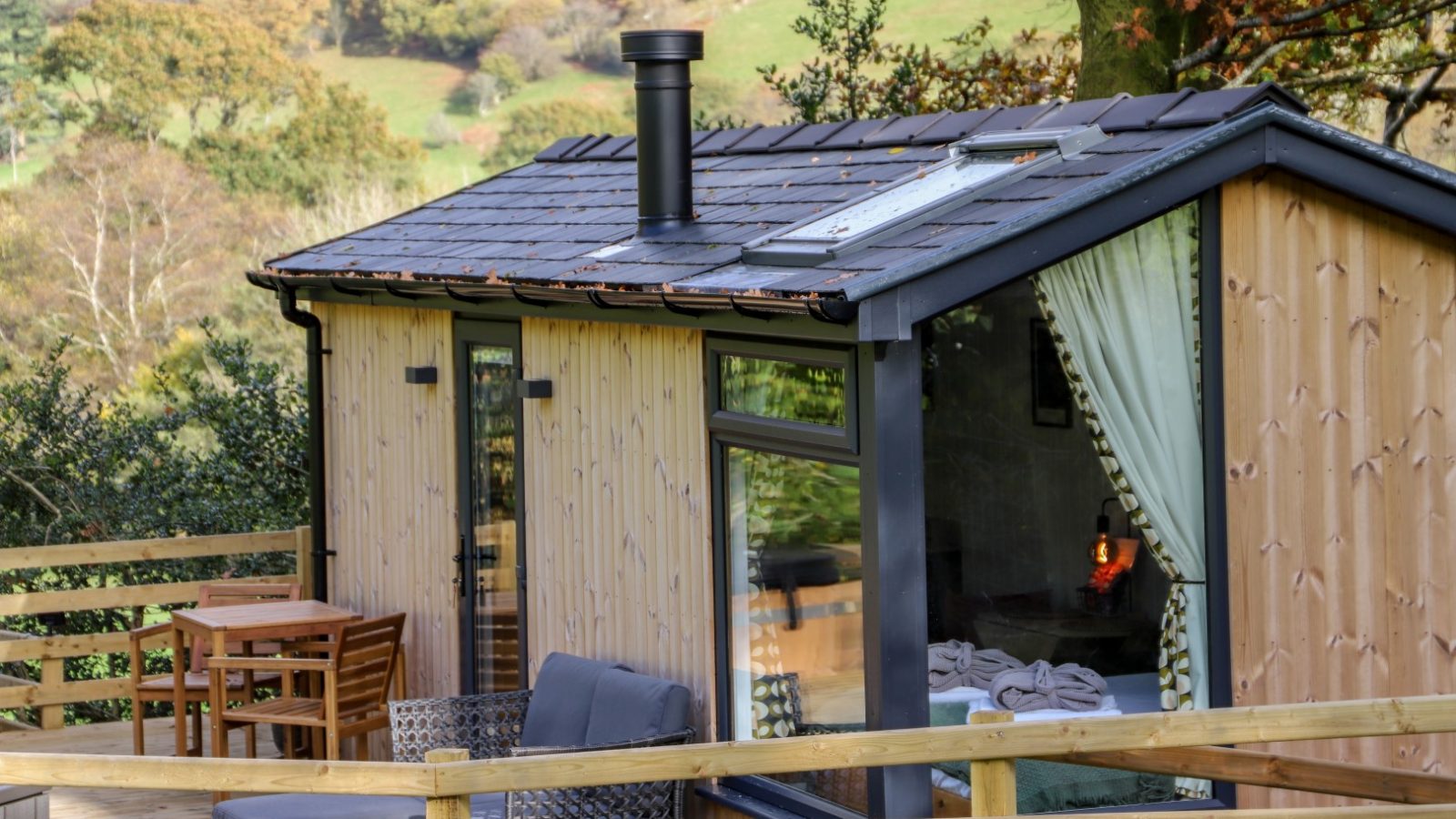 Nestled in a wooded area, this small wooden cabin features large windows and a cozy chimney. The charming deck, with chairs and a table, is perfect for enjoying your Gwyliau amidst nature's tranquility.