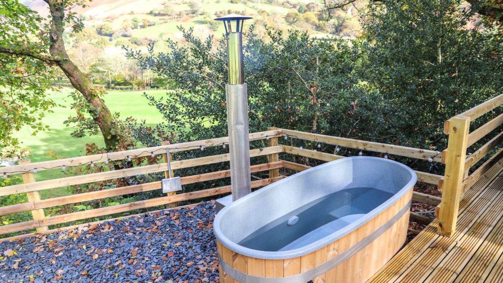 Experience the charm of Gwyliau Pennant Holidays with this outdoor wooden hot tub, featuring a metal chimney on a deck surrounded by trees. Enjoy breathtaking views of the grassy landscape and rolling hills, creating an idyllic escape.