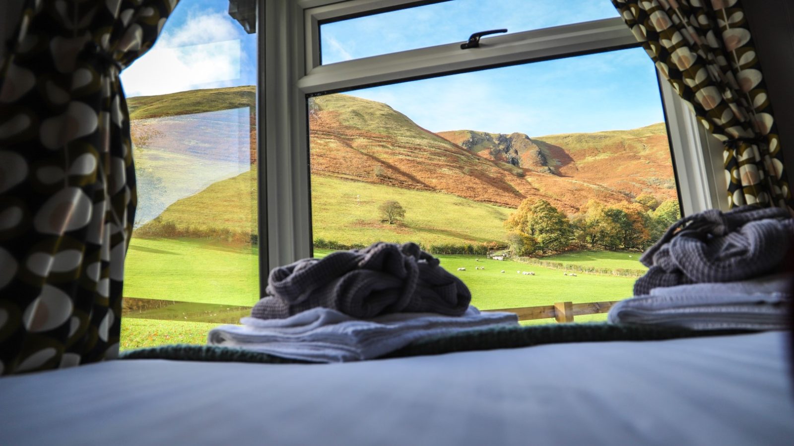 A view of rolling hills and fields through an open window, framed by patterned curtains, evokes the tranquility often sought during Pennant Holidays, with folded towels resting on the windowsill.