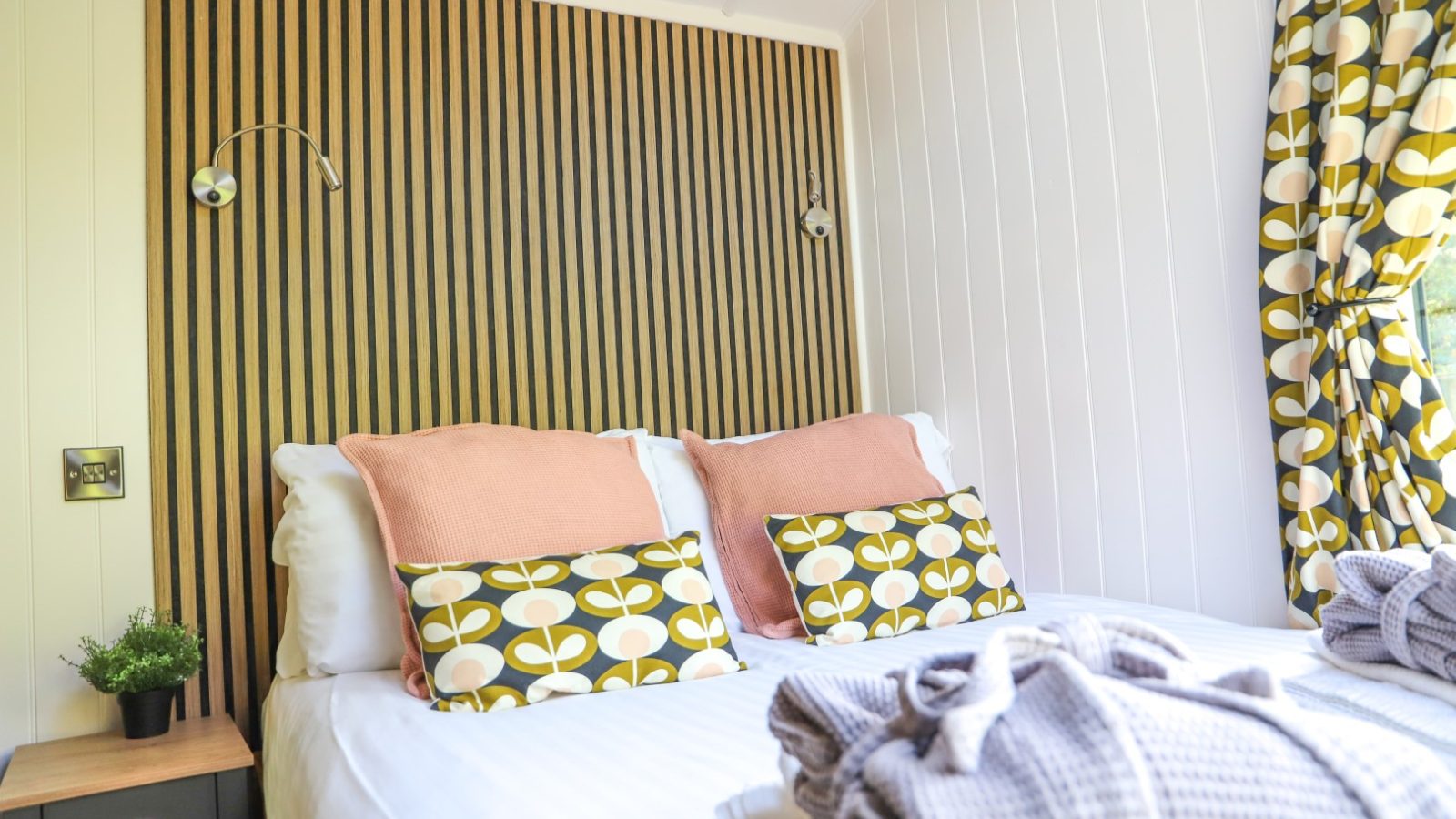 Bright bedroom with a striped wood accent wall, patterned curtains, and a neatly made bed adorned with colorful cushions and blankets, perfect for enjoying Gwyliau Pennant Holidays in comfort and style.