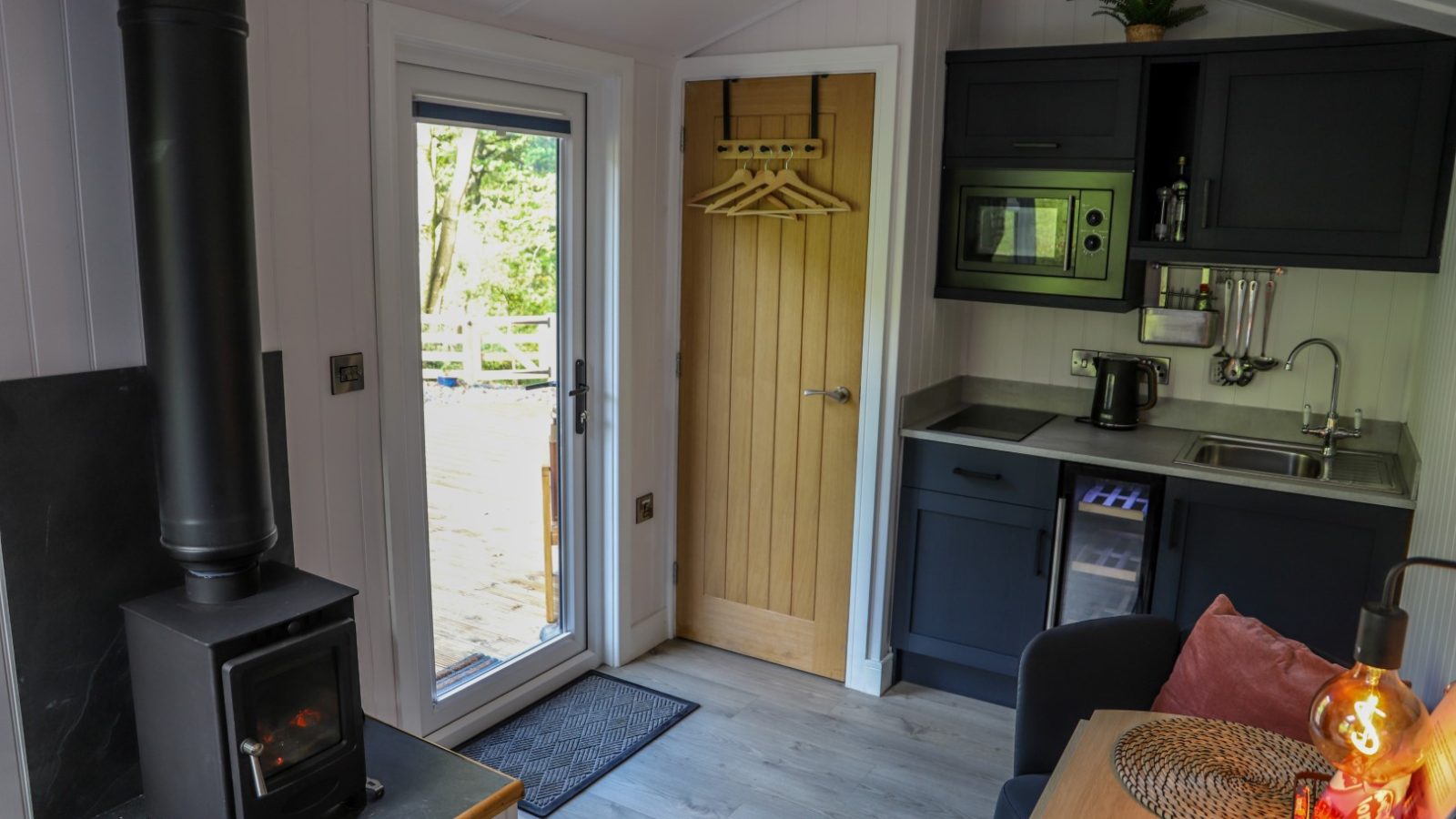 Compact cabin interior with wood-burning stove, small kitchenette, and door leading outside. Cozy and neatly organized space, perfect for your Pennant Holidays escape.