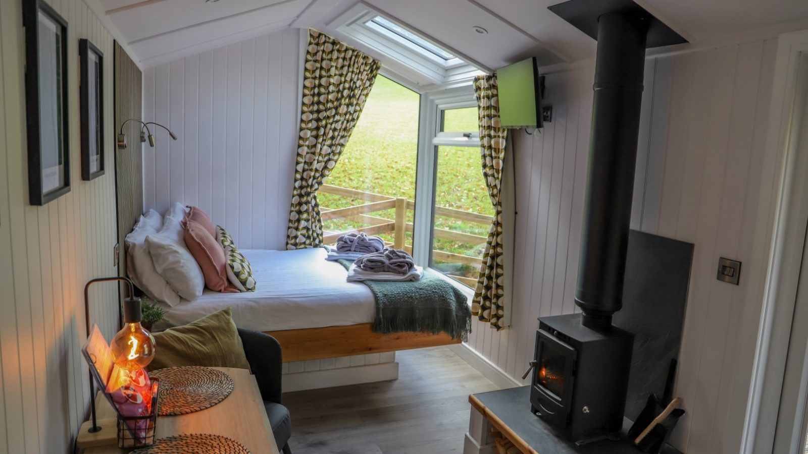 Cozy room with a bed, wood-burning stove, and a small table. A large window, adorned with patterned curtains and a cheerful pennant, overlooks lush greenery—perfect for holidays.