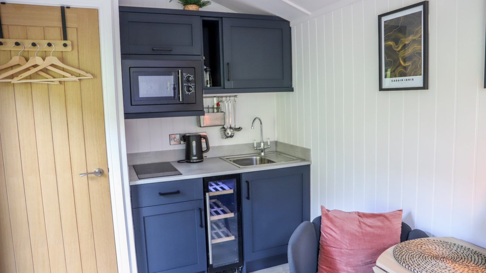 Compact kitchenette with dark cabinets, microwave, sink, and kettle. Nearby, a small table with a chair and a pink cushion—perfect for enjoying your morning coffee during your Gwyliau Pennant Holidays stay.