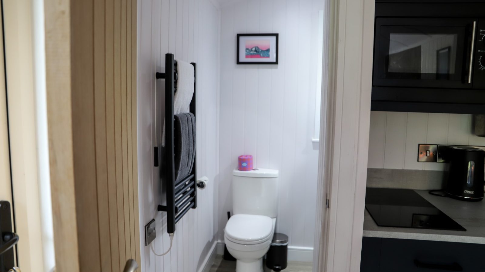 This compact bathroom features a toilet, a sleek black towel rack, wall art, and a window. Nearby, the kitchen area with visible appliances adds convenience to any stay. Enjoy your time at Gwyliau Pennant Holidays with all the essentials for a comfortable getaway.