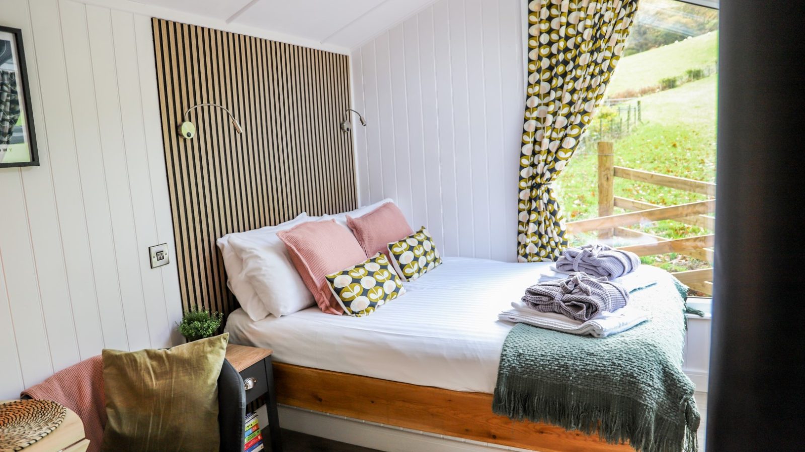 The cozy bedroom features a double bed adorned with patterned pillows and a throw blanket, complemented by a window dressed in green and white curtains. Perfect for those seeking relaxing holidays, this charming space is reminiscent of the comfort found at Gwyliau Pennant Holidays.