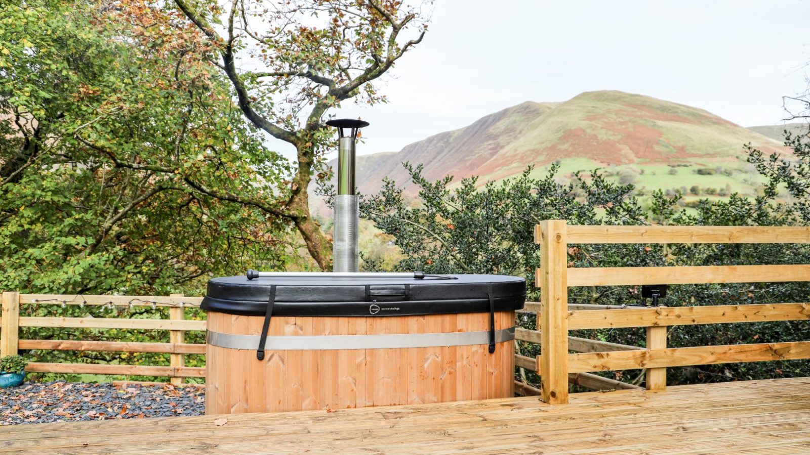 Experience a relaxing Gwyliau Pennant Holidays retreat with a wooden hot tub on the deck, offering a scenic view of rolling hills and trees in the background.