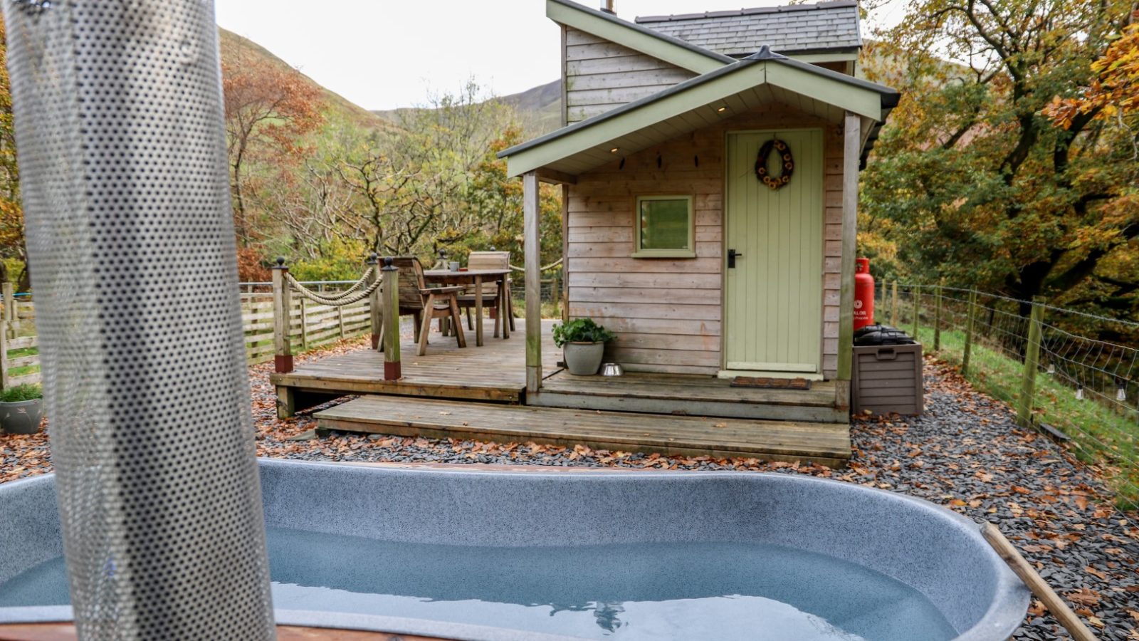 Nestled amongst the trees, this wooden cabin with a green door and porch invites relaxation. A gray outdoor tub graces the foreground, perfect for soaking in nature's tranquility. Discover the charm of Gwyliau Pennant Holidays here, where every stay feels like a magical escape.