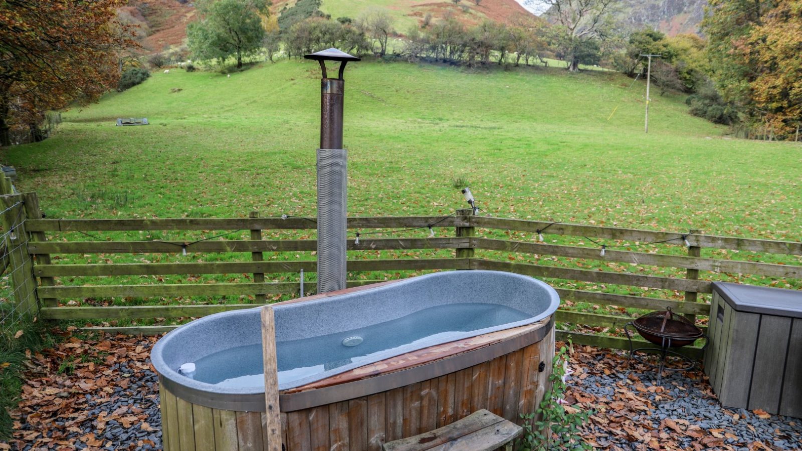 An outdoor hot tub with a chimney is set in front of a wooden fence, offering the perfect Pennant spot for holidays. It overlooks a grassy hillside adorned with vibrant autumn foliage, creating an idyllic setting for relaxation amidst nature's beauty.