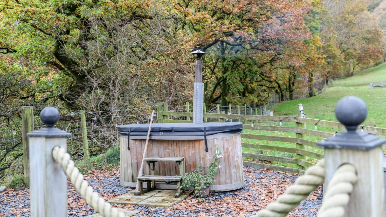 Amidst the vibrant hues of autumn leaves, a wooden hot tub with a black cover sits serenely in a fenced outdoor area, promising relaxation and rejuvenation—the perfect retreat to unwind during your Gwyliau Pennant Holidays.