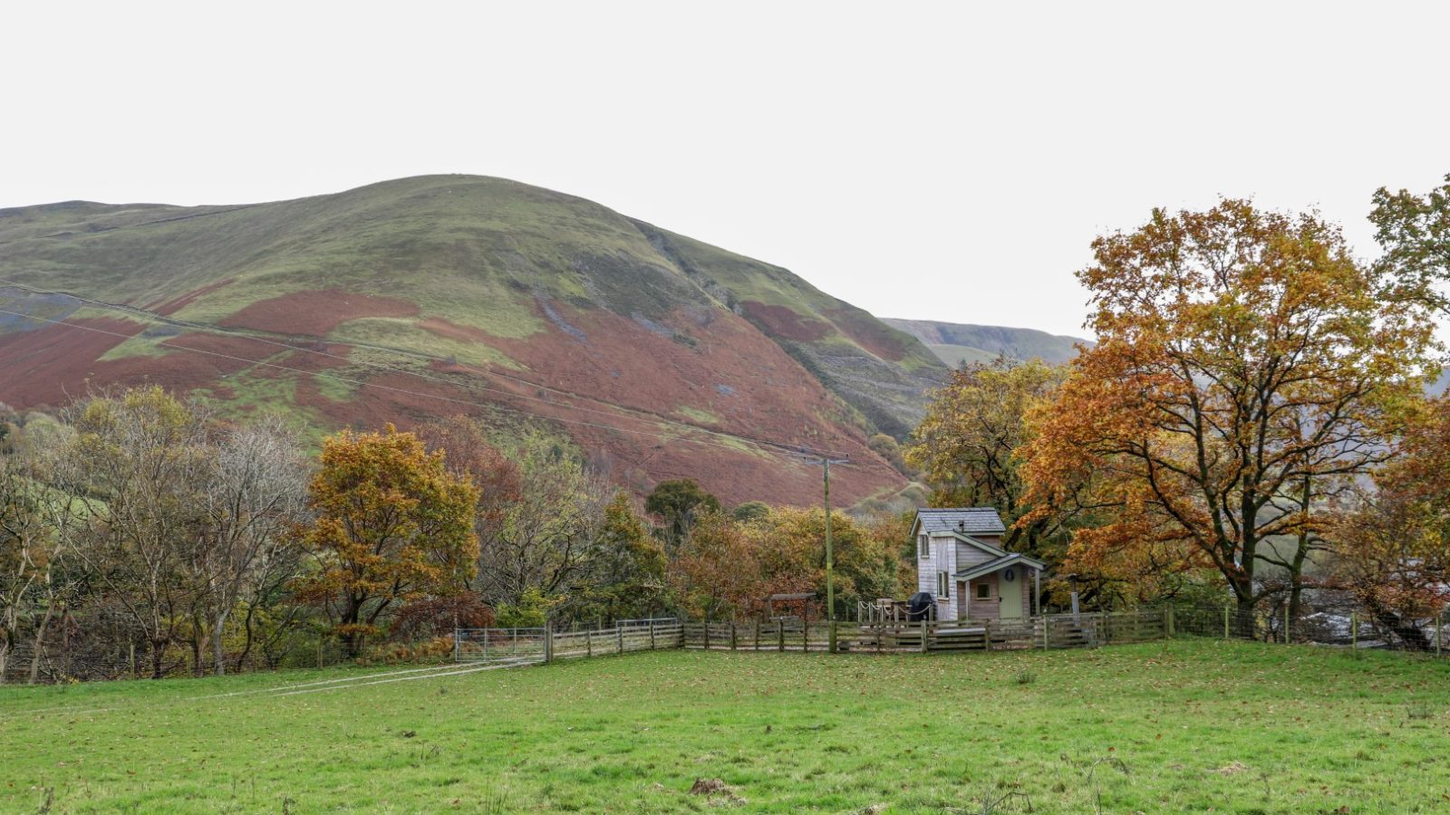 Nestled against rolling hills and an overcast sky, this small Gwyliau Pennant Holidays house boasts a fenced yard surrounded by lush trees and greenery.