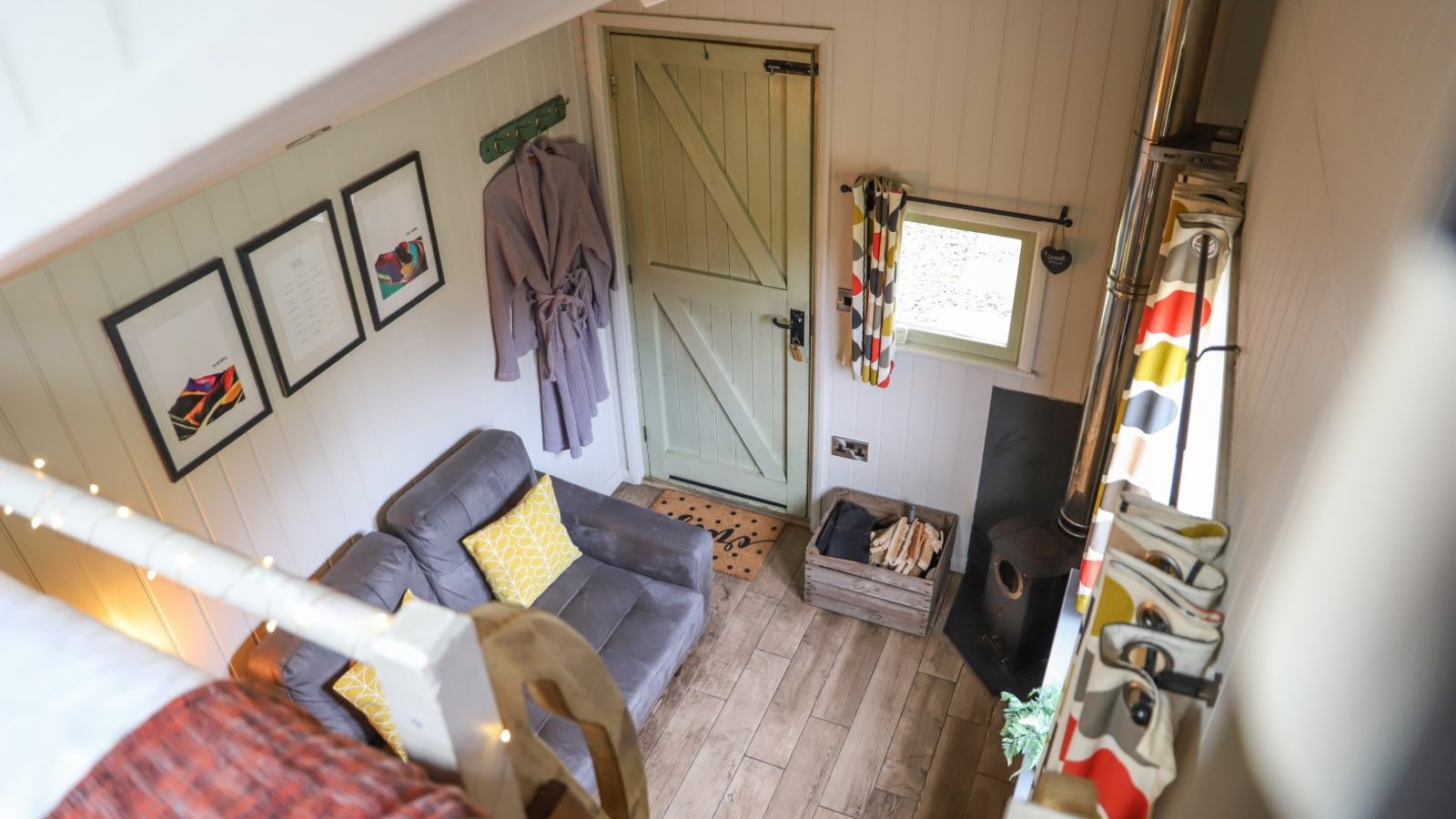 Cozy room with wood flooring, a couch, and a green door, this charming space by Gwyliau Pennant Holidays features framed art, a robe on hook, colorful curtains, and a wood stove with logs.