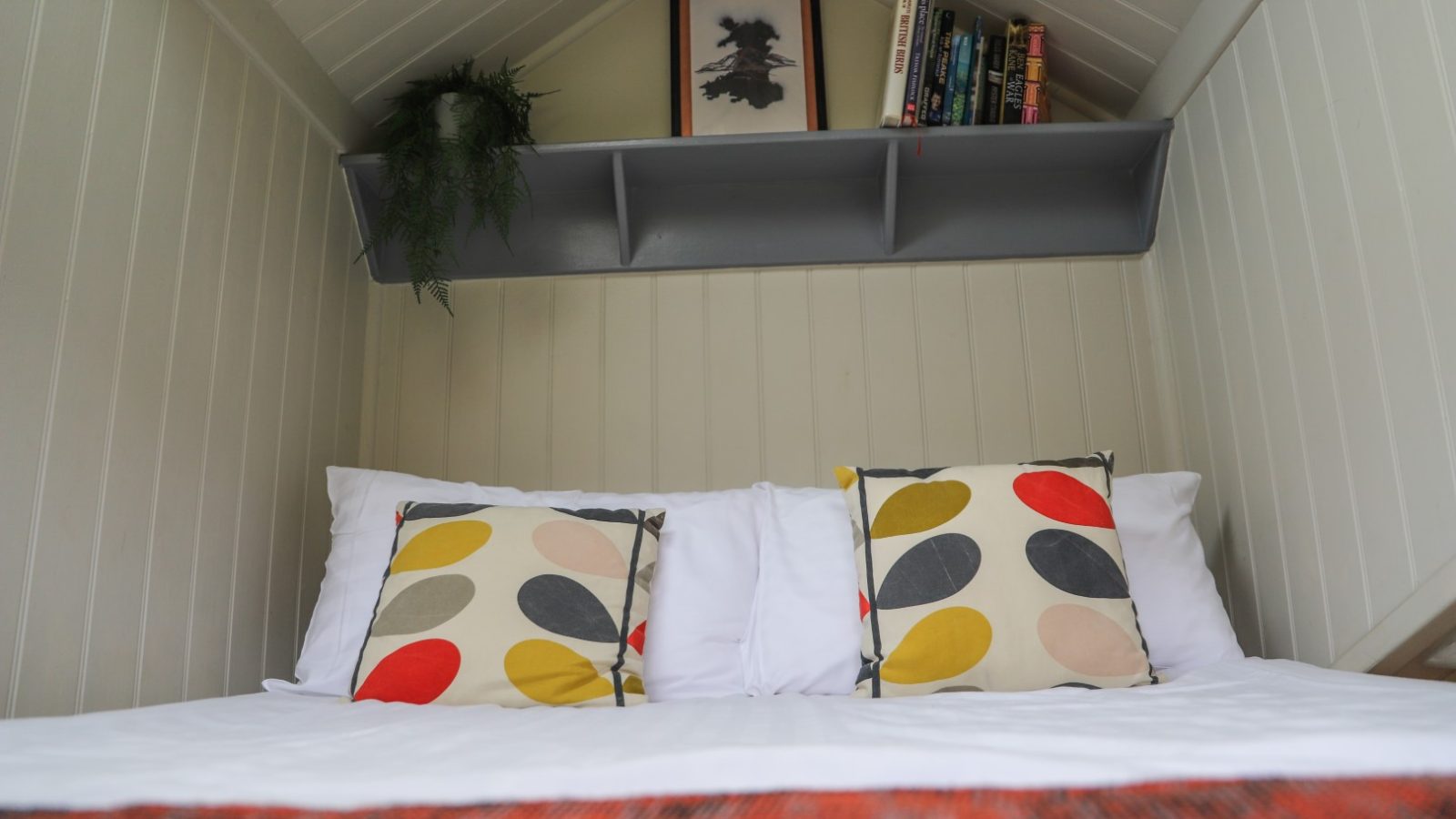 Cozy bedroom with a bed featuring two patterned pillows, a red blanket, and a shelf above holding books and decor items, creating the perfect retreat for Gwyliau Pennant Holidays.