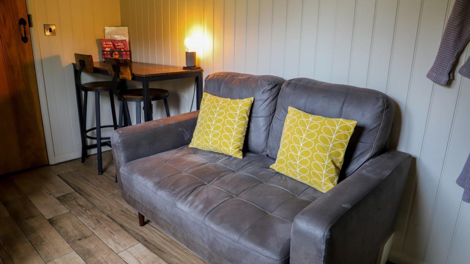 Cozy living room with a gray sofa and two yellow pillows, a small table with chairs, wooden floor, and soft ambient lighting—a perfect retreat that echoes the warmth of Gwyliau Pennant Holidays.