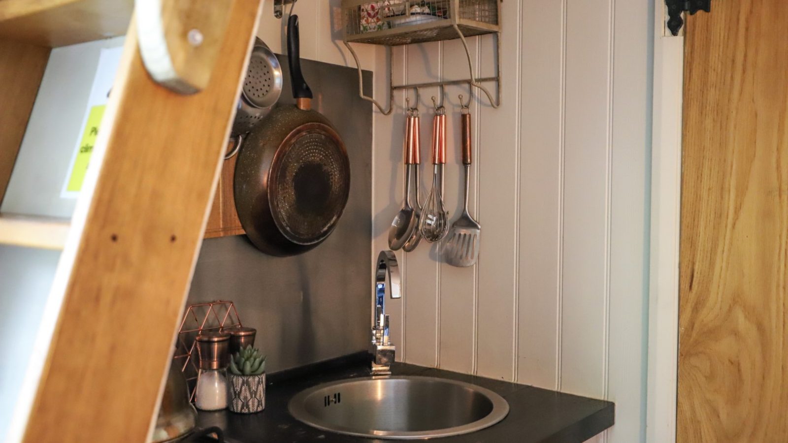 Compact kitchen with a round sink, utensils hanging on the wall, and pots on a shelf. A wooden ladder is nearby, adorned with a festive pennant that adds a touch of holiday charm.