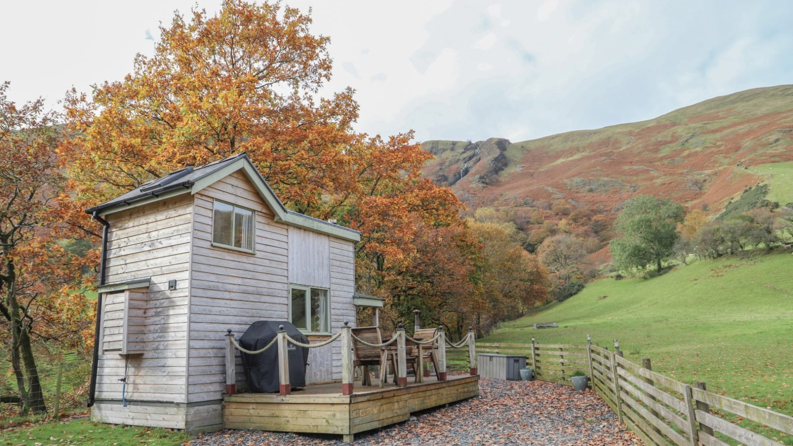 Cozy small wooden cabin with a deck, set among autumn trees near rolling hills and a fence-lined path under a partly cloudy sky—a perfect retreat for Gwyliau Pennant Holidays.