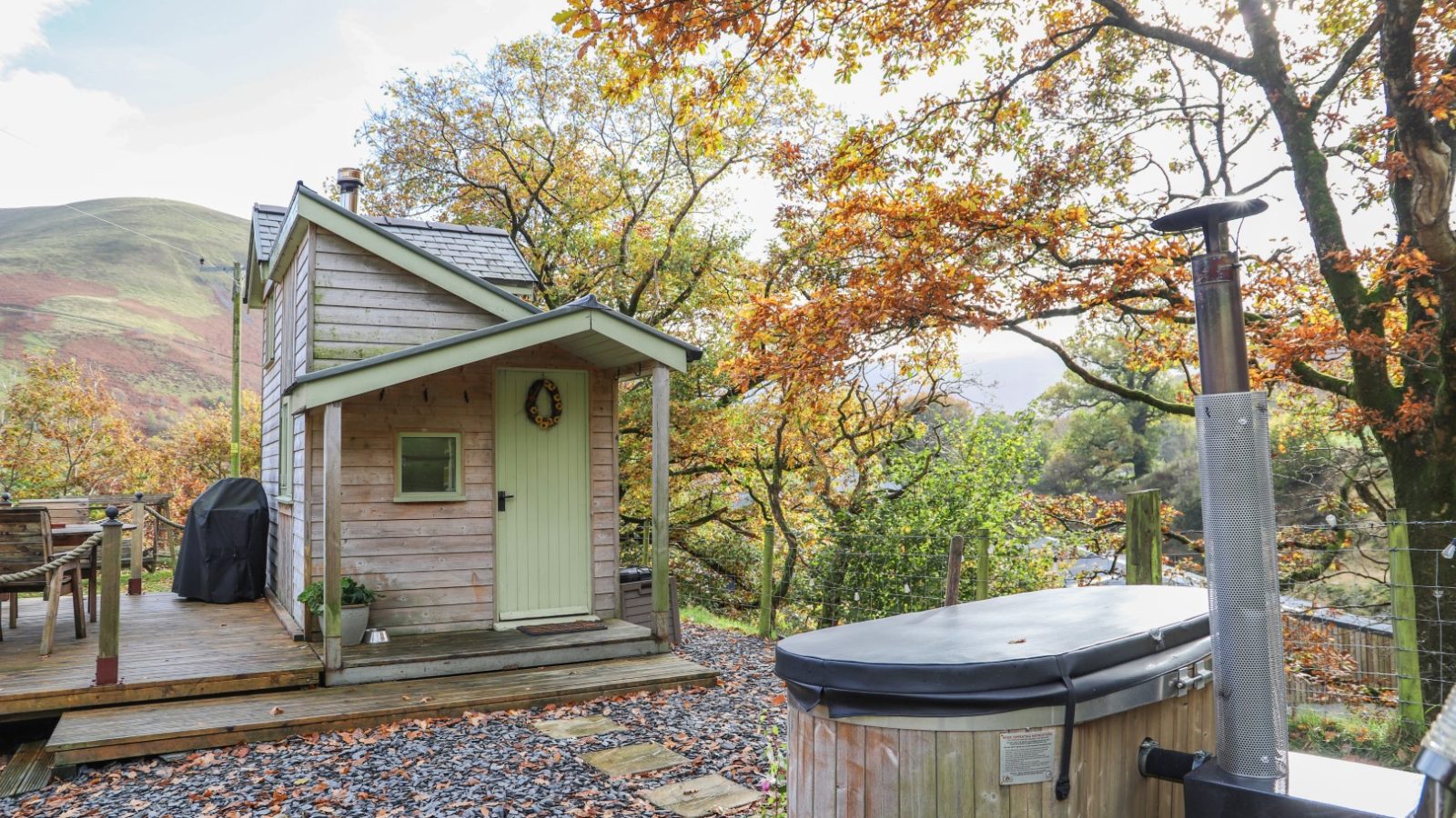 A cozy wooden cabin nestled among autumn trees offers a perfect retreat. With a deck and hot tub in the foreground, it's an idyllic escape for memorable gwyliau holidays.