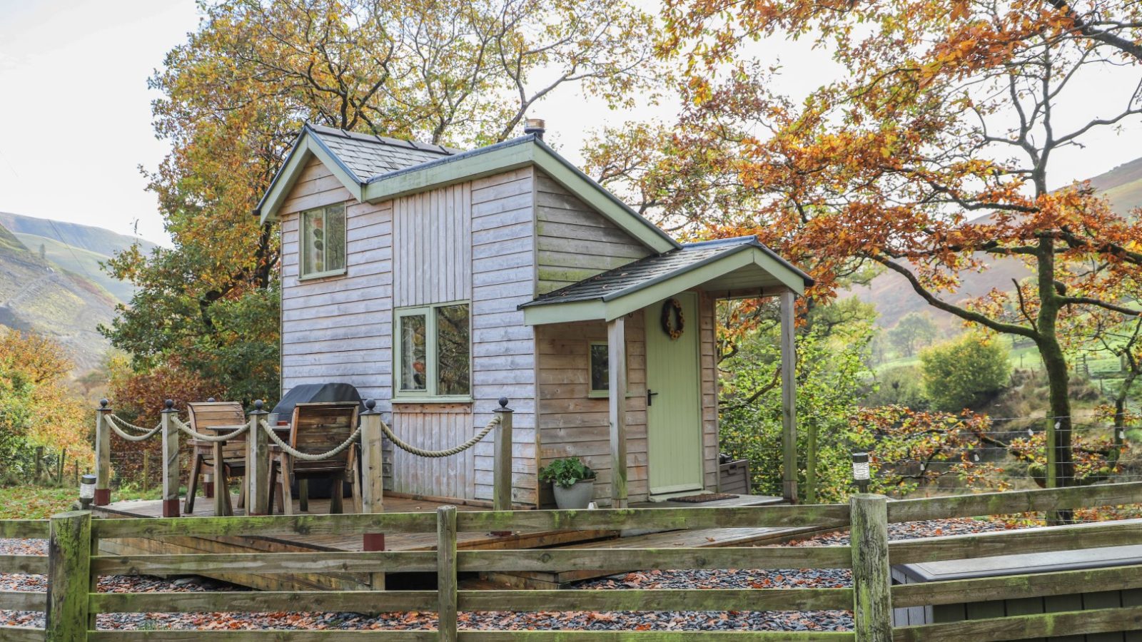 A small wooden cabin with a deck is nestled in an autumn forest, surrounded by colorful trees and hills, perfect for Gwyliau Pennant Holidays.