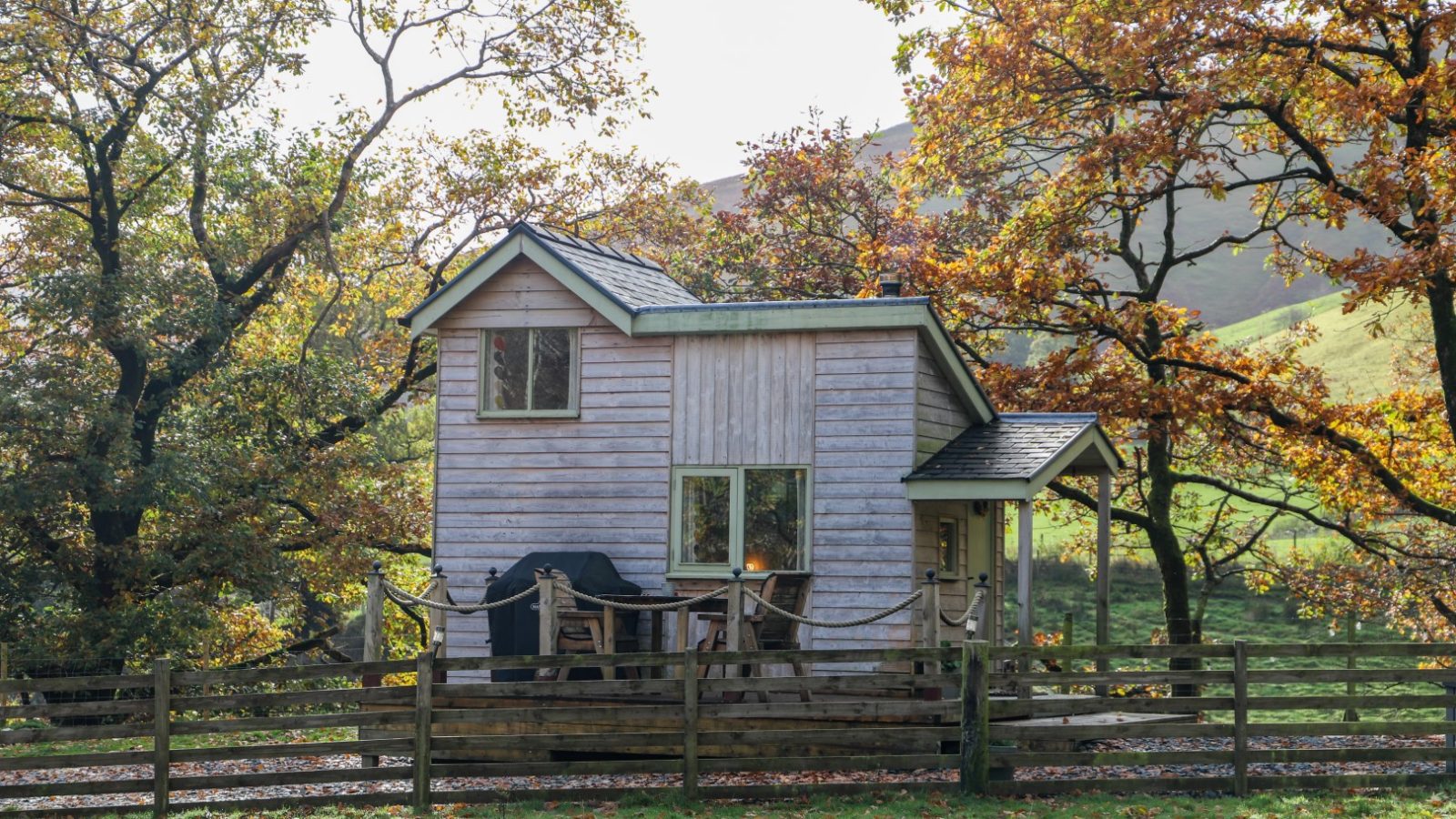 A small wooden cabin, perfect for holidays, is nestled among autumn trees. A wooden fence stands in the foreground, while hills create a stunning backdrop.