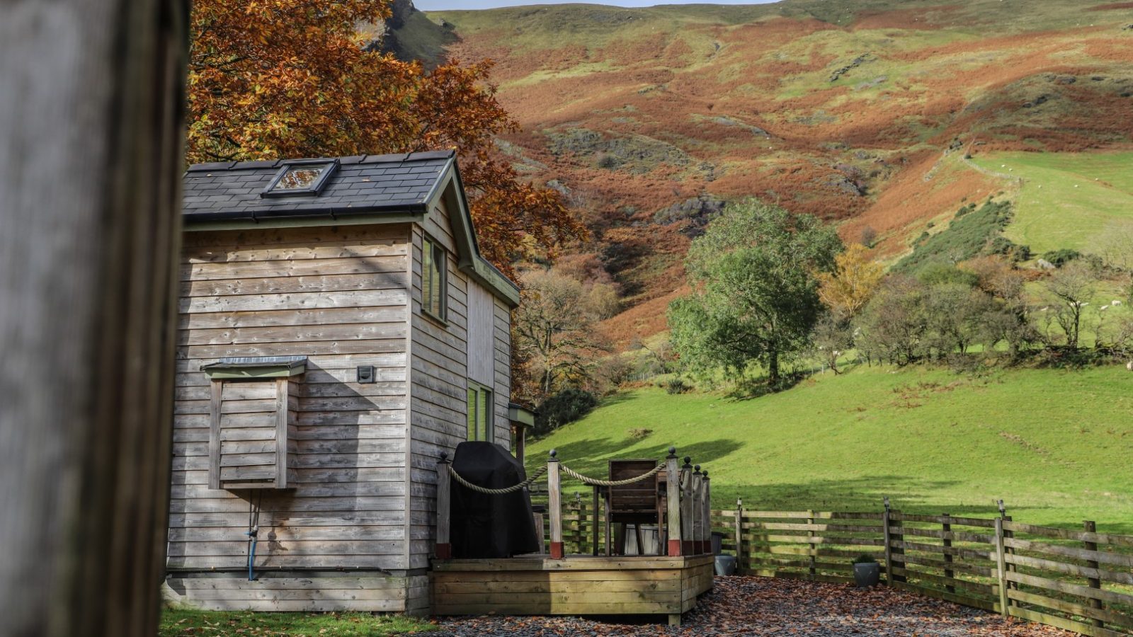 Tiny wooden cabin with a deck, surrounded by autumn trees and green hills, under a partly cloudy sky—a perfect escape for Gwyliau.