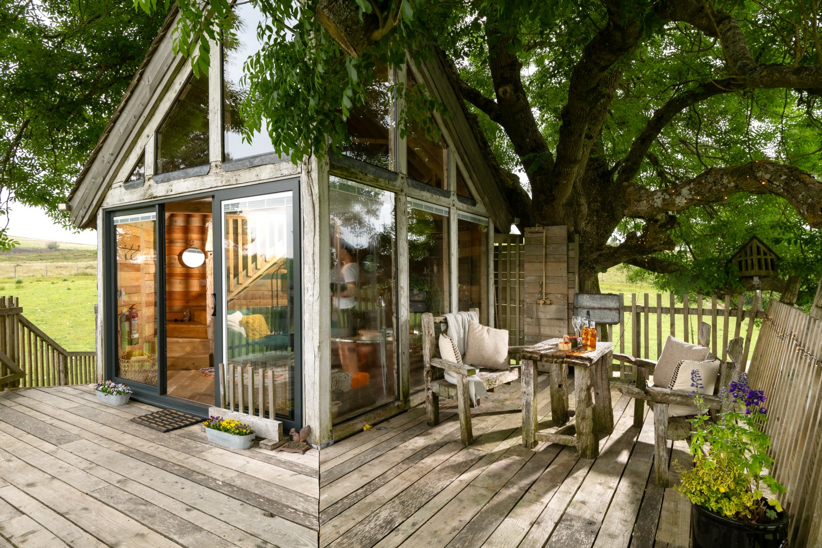 Treehouse with glass walls and a wooden deck featuring rustic furniture, set amidst lush greenery with a breathtaking view of the countryside—a perfect escape. Enter our giveaway for a chance to win a serene getaway in this idyllic retreat, straight from your landing page dreams.