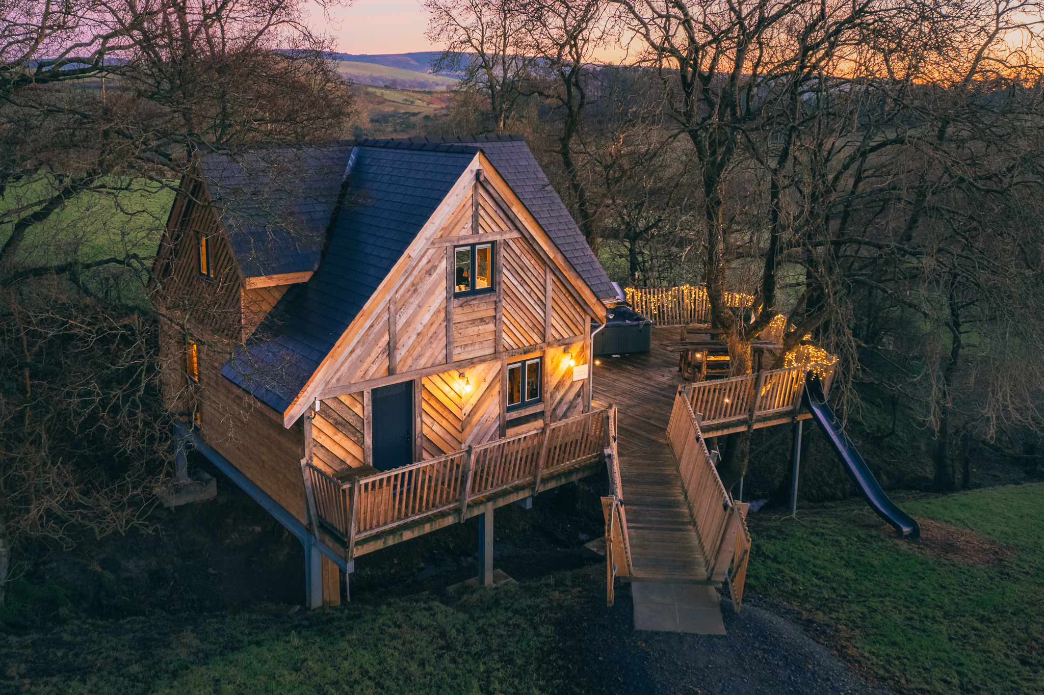 A wooden treehouse with a porch and slide, elevated on stilts, serves as the perfect backdrop for our exciting giveaway nestled among the trees at sunset in a rural landscape.