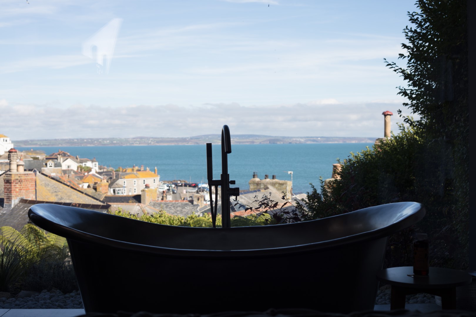 Silhouette of a bathtub in front of a large window overlooking the serene beauty of a coastal Cornwall town, with sweeping ocean views under the expansive blue sky.