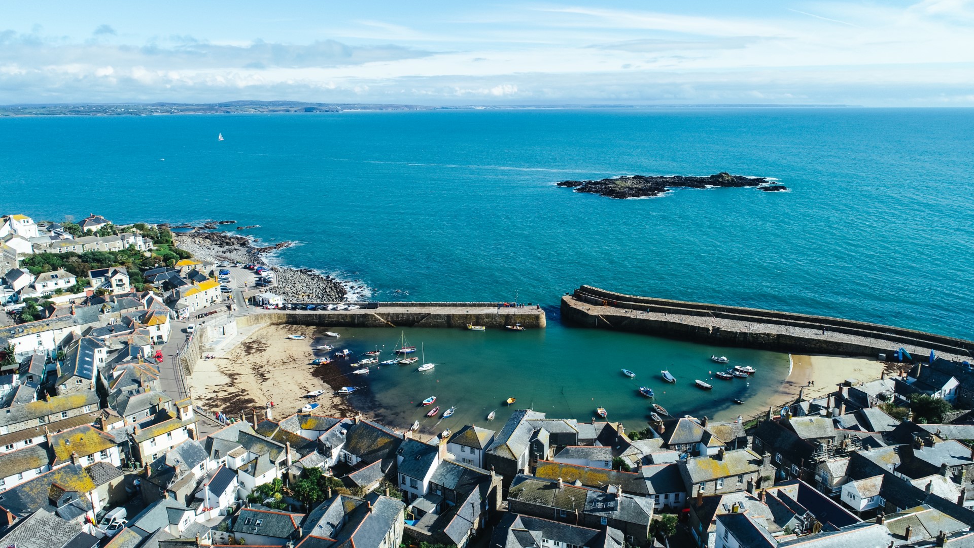 Aerial view of a coastal Cornish town in the UK with boats nestled in a small harbor and a breakwater stretching into the blue sea.