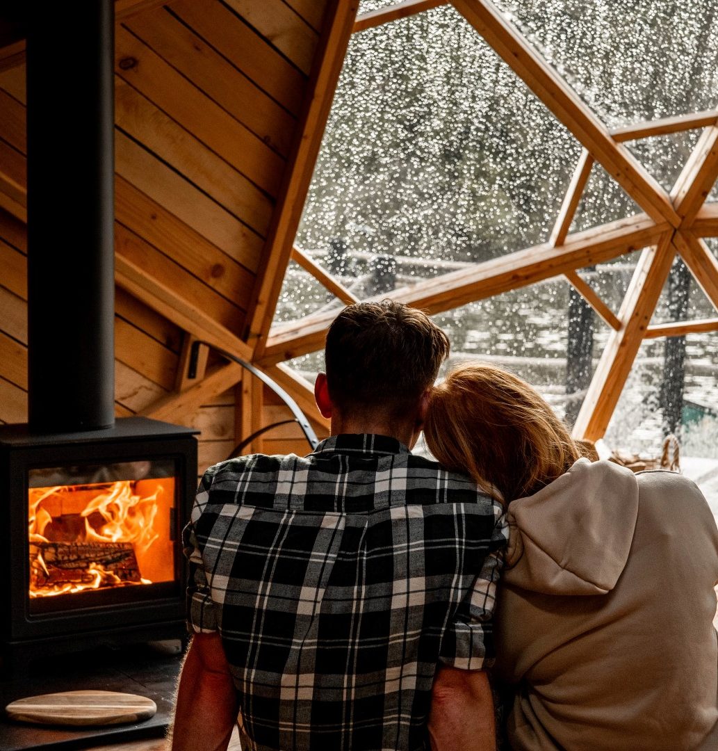 In the heart of a cosy cabin, a couple sits by a wood-burning stove. The warmth of home surrounds them as they gaze through a large geometric window, admiring the snowy scenery outside. The perfect accommodation for a romantic getaway for two