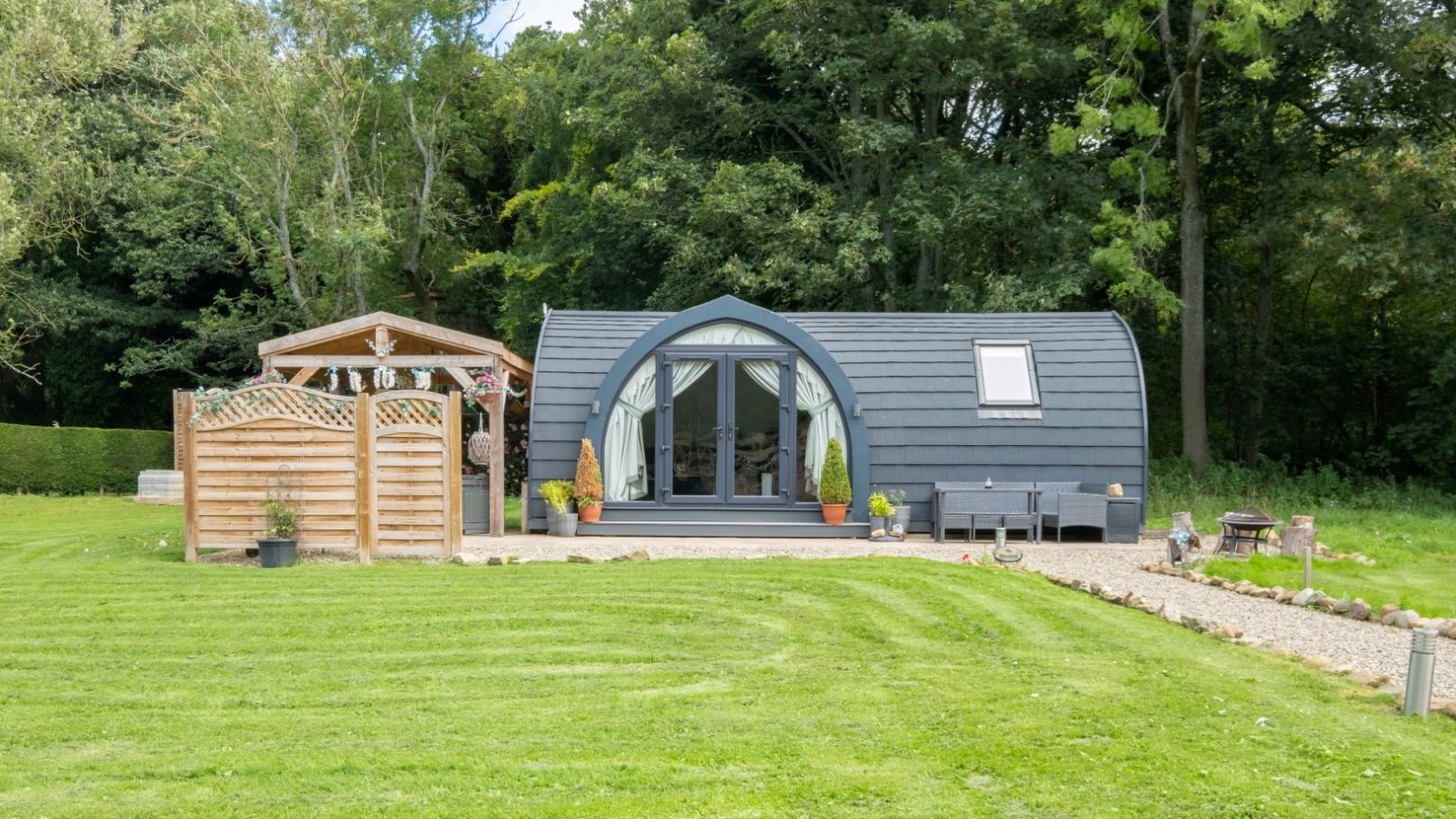 A small, arched-roof glamping cabin with a glass front door and patio in a grassy yard, surrounded by trees offers the perfect retreat.