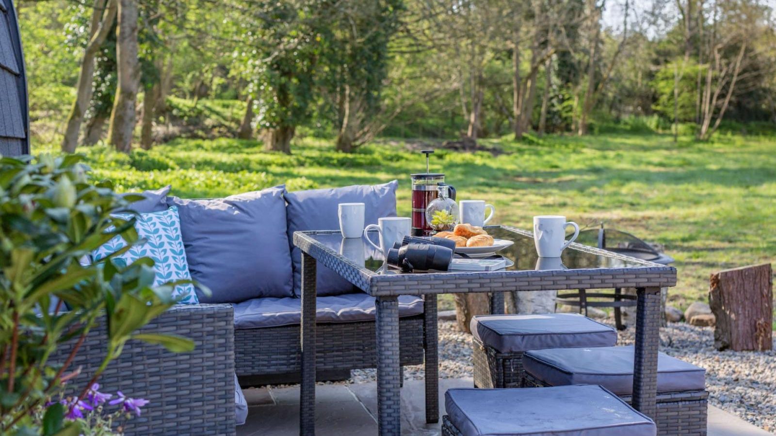 Discover a hidden retreat on your outdoor patio setup, featuring a glass table, cushioned seating, a coffee pot, mugs, croissants, and an expansive view of the lush green landscape. Perfect for those who yearn for a touch of glamping right at home.