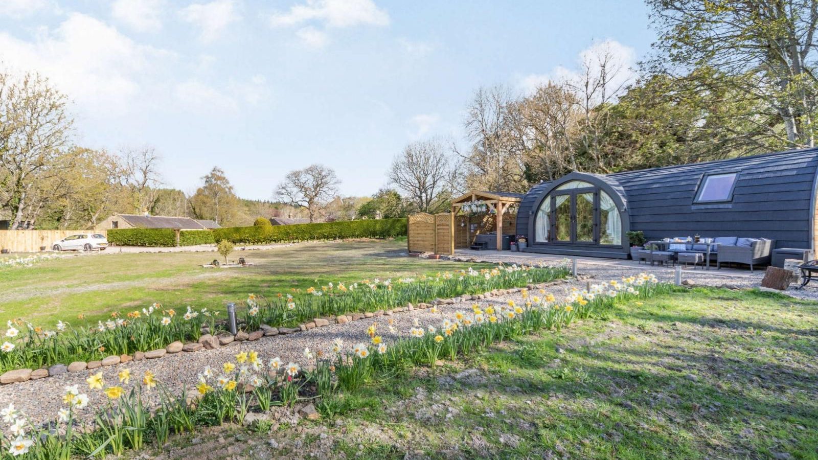 Modern cabin with arched window, outdoor seating, and a lawn bordered by yellow flowers under a clear blue sky; the perfect hidden retreat.